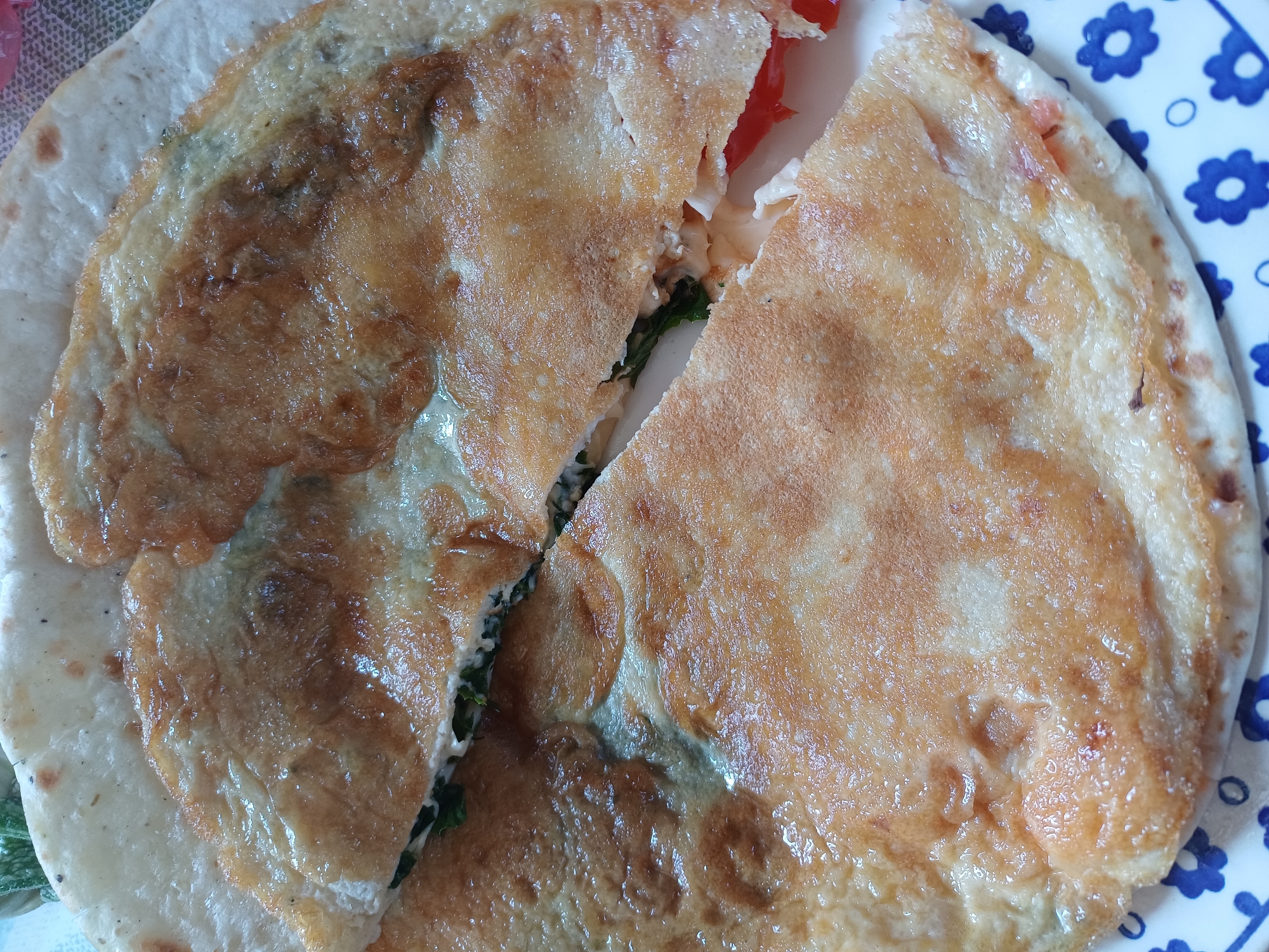A close-up, overhead shot captures a partially eaten flatbread dish, likely a savory crepe or thin pancake, served on a plate with a blue and white floral pattern. The dish, which appears to be cut into two halves, is filled with green herbs, possibly spinach or parsley, and pieces of red tomato. The flatbread itself is golden brown and slightly oily, with some darker, crisped edges. The background is blurred, but hints of a placemat or tablecloth are visible. The image is taken from a high angle, focusing on the food item and its presentation.