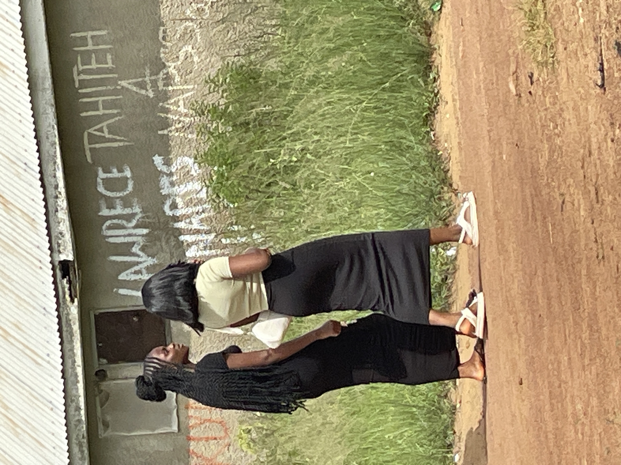 Two young women are standing on a dirt road in front of a building with a corrugated metal roof. The building has a rough, textured wall with white graffiti on it. The graffiti appears to say "LAWRENCE TAHITEH A NAPES VAPSS". The woman on the left has long braided hair and is wearing a black dress and sandals. She is holding a white handbag. The woman on the right has dark hair and is wearing a light-colored t-shirt and a long black skirt with white sandals. She is facing away from the camera. Tall green grass and plants are growing behind the women and in front of the building. The ground is a reddish-brown dirt. The scene appears to be in Durumi, Nigeria, based on the style of the building and the surrounding vegetation.