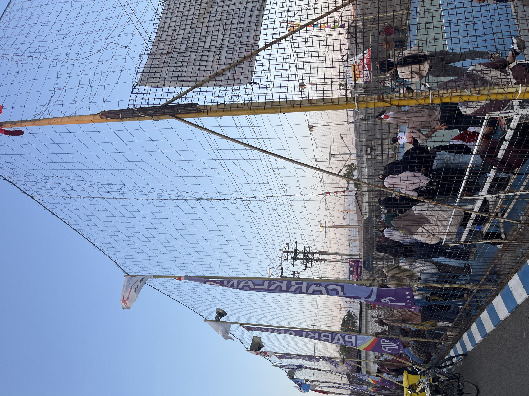 The image depicts an outdoor volleyball court in the city of Pelang, Indonesia. A large net divides the court, with a blue surface. Several flags, primarily purple with white lettering, are displayed along the fence surrounding the court. A white flag with red and blue markings is also visible. A group of people are gathered behind the fence, some watching the game on the court and others conversing. A bicycle is parked near the fence on the left side of the image. The sky is clear and blue, suggesting it is daytime. In the background, there are buildings and electrical poles.