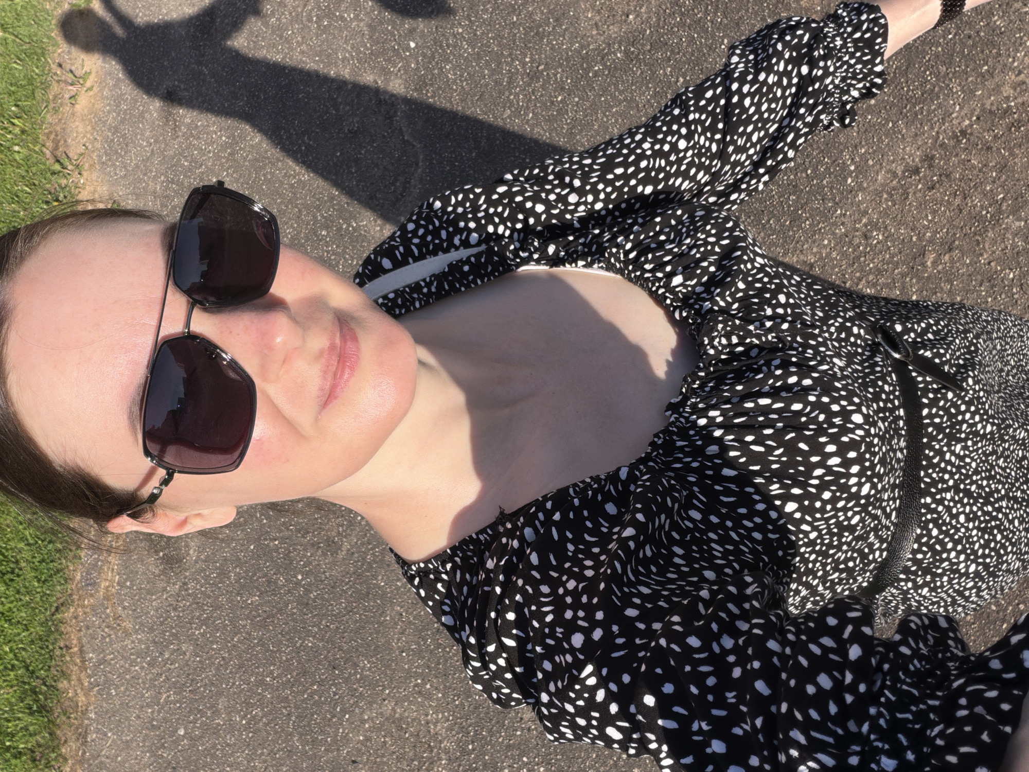 The image shows a woman wearing sunglasses and a black and white patterned dress. She is standing outdoors on a paved surface next to a patch of grass. Her shadow is visible in the background.