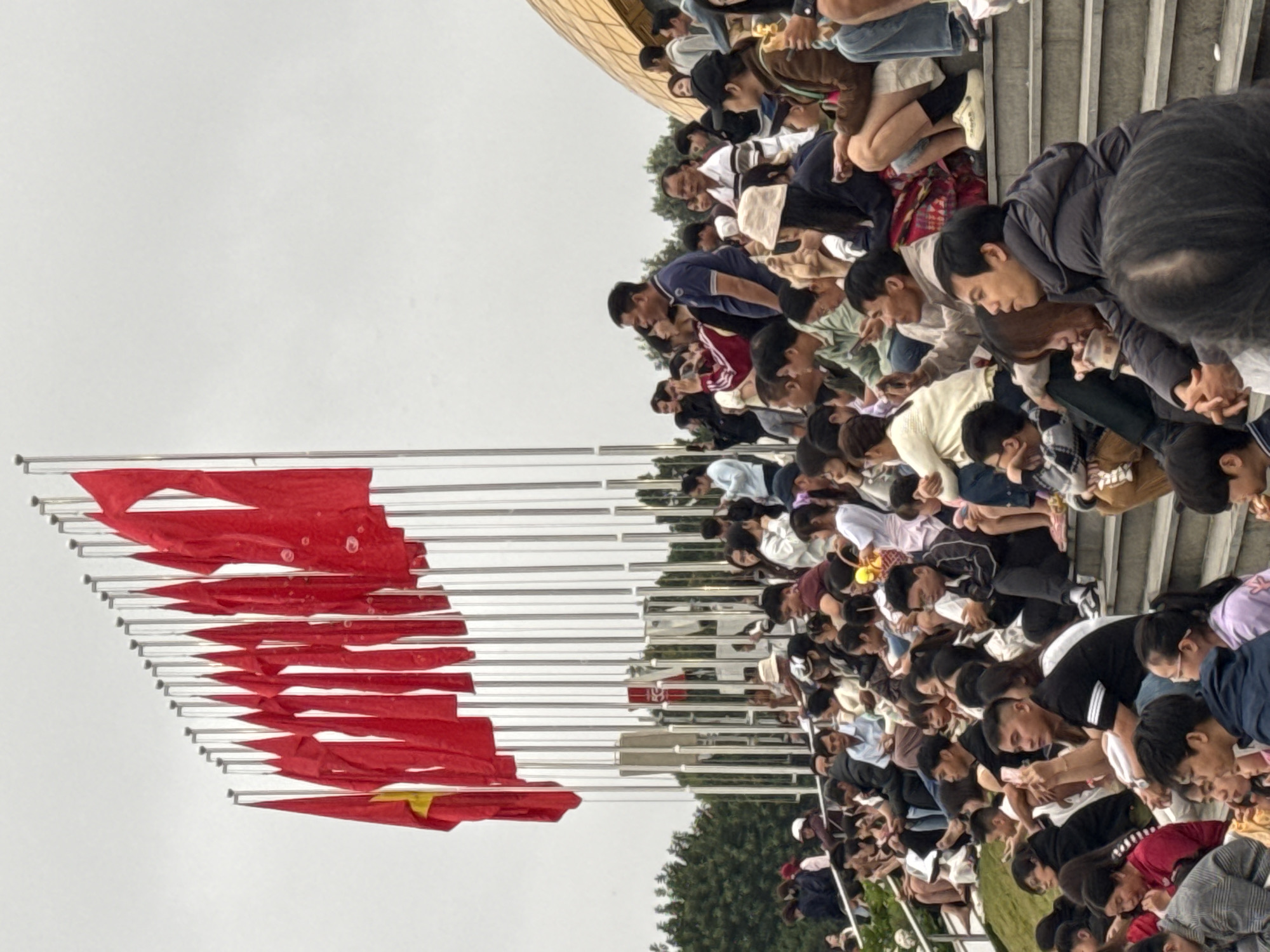 A wide shot captures a crowd of people seated on tiered concrete steps outdoors, facing a display of numerous red flags on poles. The flags, some with yellow stars, are arranged in a long row and appear to be waving gently. The background includes a modern building with a golden, curved dome and some trees. The sky is overcast, suggesting a cloudy day. The people are diverse in age and are dressed in casual clothing. Some are looking towards the flags, while others are engaged in conversations or using their phones. The overall scene suggests an outdoor event or gathering in Da Lat, Vietnam.