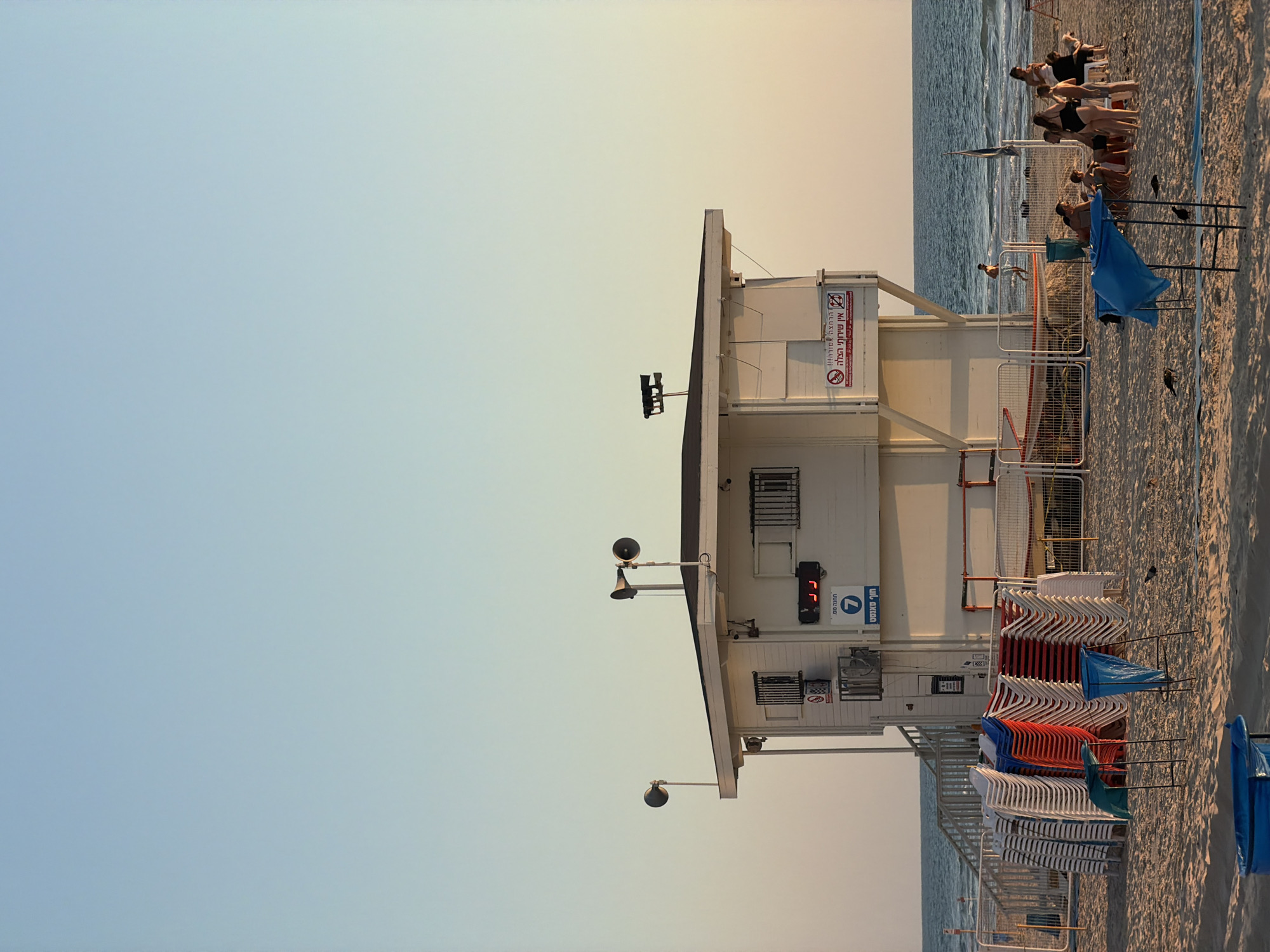 The image depicts a wide shot of a beach in Bat Yam, Israel, under a clear sky transitioning from light blue to a pale yellow near the horizon. In the center of the frame stands a two-story white lifeguard station with a dark roof. It has windows with metal bars and is equipped with two large speakers on poles. A digital clock with red numerals is visible on the side of the building, and a sign with Hebrew text and a number "7" is also present.
In front of the lifeguard station, numerous stacks of colorful plastic chairs (white, red, and blue) are neatly arranged on the sand. Several fences delineate areas on the beach, and a set of stairs leads up to the lifeguard station.
To the right of the lifeguard station, a group of people are relaxing on the sand, some sitting, some standing. They are dressed in beach attire. A blue tarp is spread over a structure in the foreground, and a few birds are visible on the sand. The ocean stretches across the background, with gentle waves reaching the shore. The overall scene is one of a typical beach day in Bat Yam, with infrastructure for beach safety and recreation.