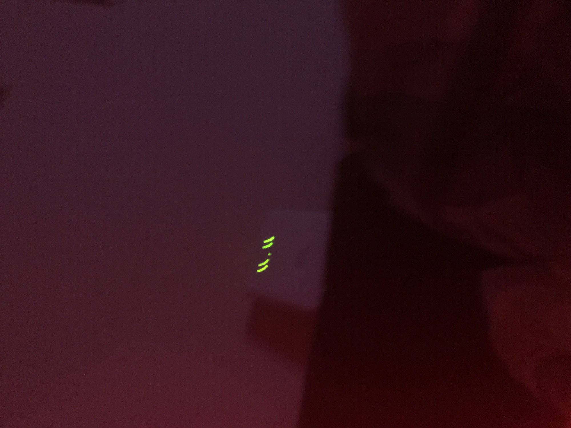 A dimly lit, indoor shot features a dark, abstract background with a wall or surface on the left side, displaying a red-purple hue. Three rows of short, horizontal, green lights are visible on a white or light-colored object. To the right, a darker, shadowy area suggests the presence of a person, with a blurred face or head partly visible. There's no clear evidence of a location in Yerevan, Armenia. No specific actions or interactions are depicted.
