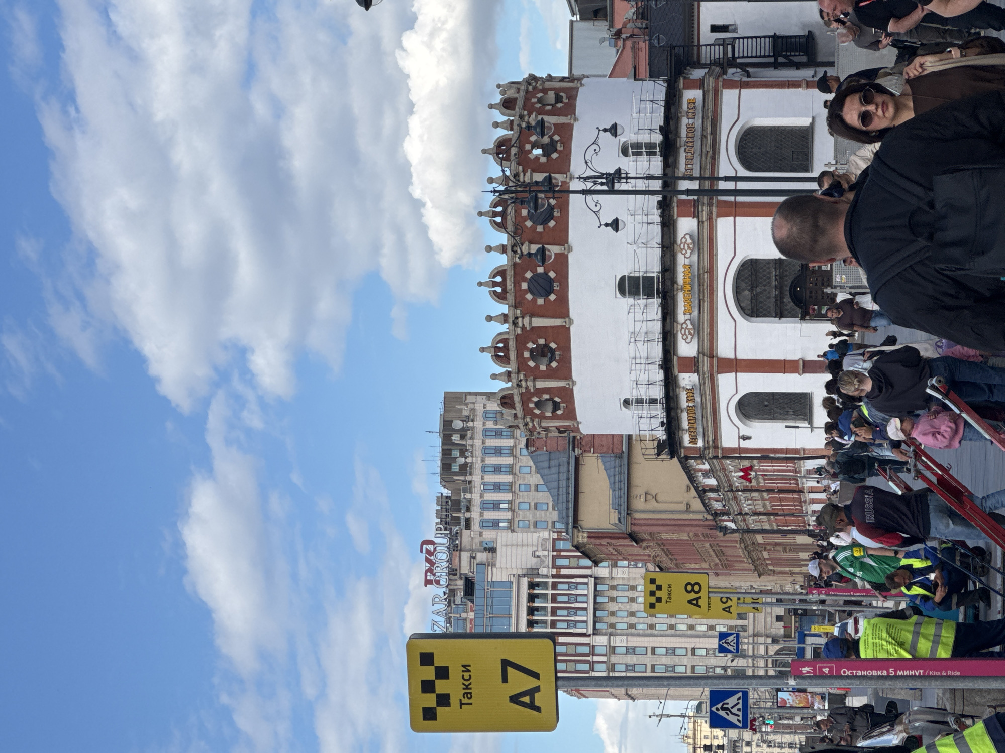In Moscow, Russia, on a day with a partly cloudy sky, people are gathered on a city street. A yellow taxi sign with "Такси" and "A7" is prominently displayed on the left side of the image. In the background, modern and older buildings are visible, including a tall building with "ZAR GROUP" signage at the top. A distinct circular building with red and white decorative elements and arched windows is also present. A street lamp with multiple lights stands near this building. A crowd of people, some wearing safety vests, are walking and standing on the street. Some are looking at their phones, while others appear to be interacting. Pedestrian crossing signs are visible on poles.