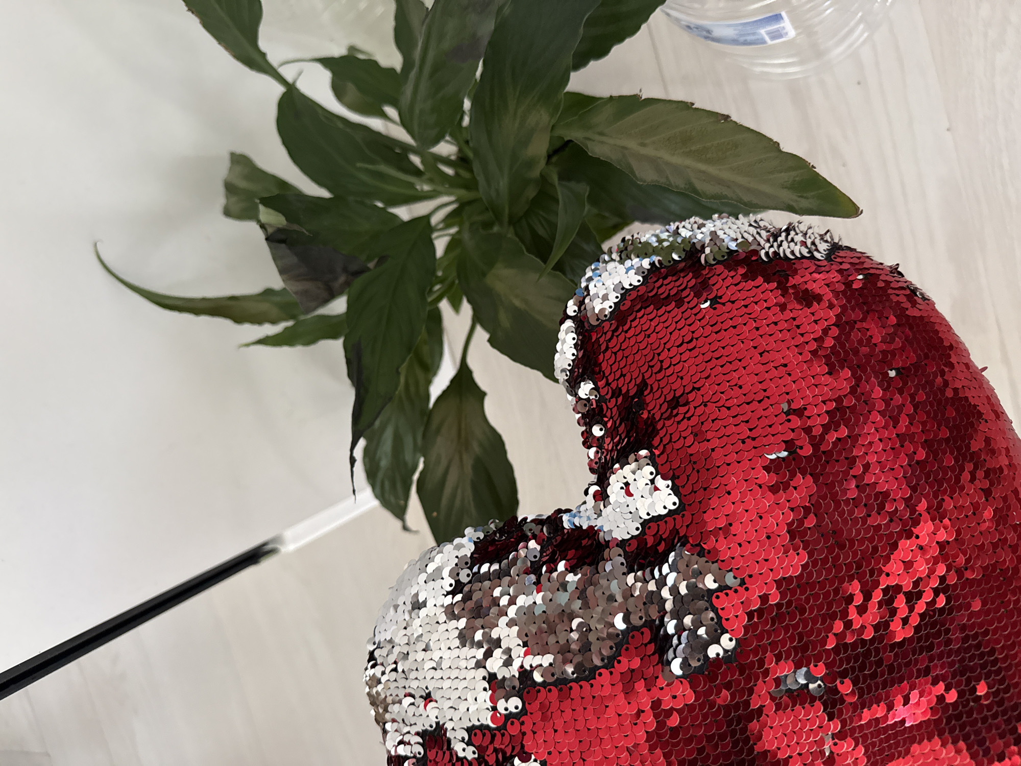 A close-up, eye-level shot captures a red heart-shaped pillow adorned with reversible sequins, alternating between red and silver. The pillow is positioned on a light-colored wooden floor. To the right of the pillow, a lush green plant with broad leaves sits in a pot, partially obscuring a clear plastic bottle. In the background, a white wall is visible, with a black and clear object, possibly part of a lamp or stand, extending from the upper left corner. The scene appears to be set indoors, potentially in a residential or commercial space in Sofia, Bulgaria, though no specific location markers are visible.