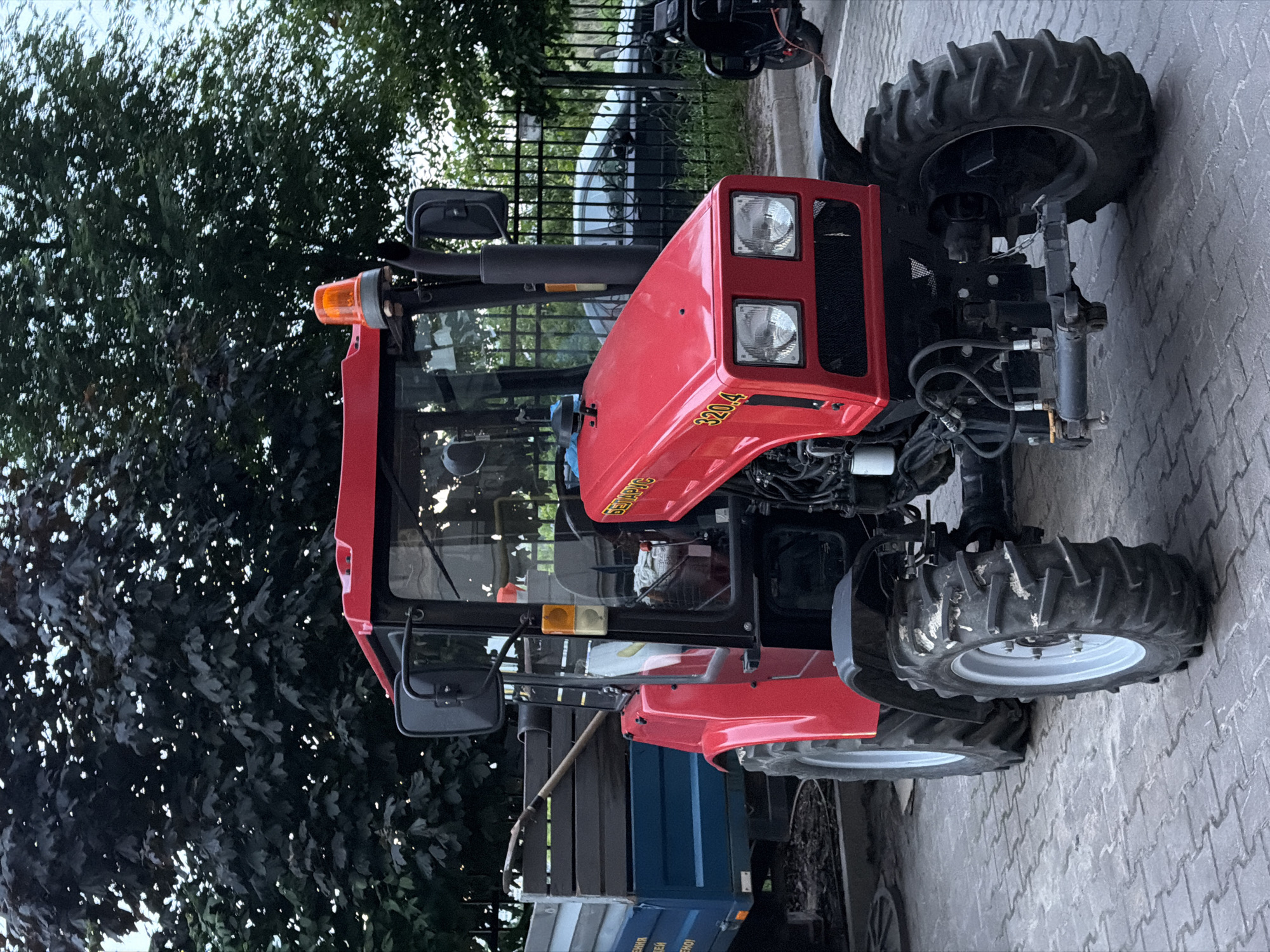 A red tractor is prominently featured, parked outdoors on a paved surface. The tractor is painted red with black and gray components, including its cab frame and exhaust pipe. It has large, deeply treaded tires. The front of the tractor displays two rectangular headlight clusters, each with two lights. An orange beacon light is mounted on the left side of the cab roof.
Visible text on the side of the engine hood reads "BELARUS" in white letters with a yellow outline, and "320.4" in yellow numerals. Another yellow inscription, possibly "MTZ" or a similar Cyrillic abbreviation, is also present.
The tractor is stationary, and no people are visible in the image. The background features dense green foliage from trees, suggesting a natural environment. Behind the tractor, a black metal fence separates it from a partially visible vehicle and possibly other structures. A blue trailer or container is attached to the rear of the tractor. The ground is paved with light gray pavers. The lighting indicates it is daytime, possibly with overcast conditions or during the late afternoon. The scene takes place in Sofiivska Borshchahivka, Ukraine.