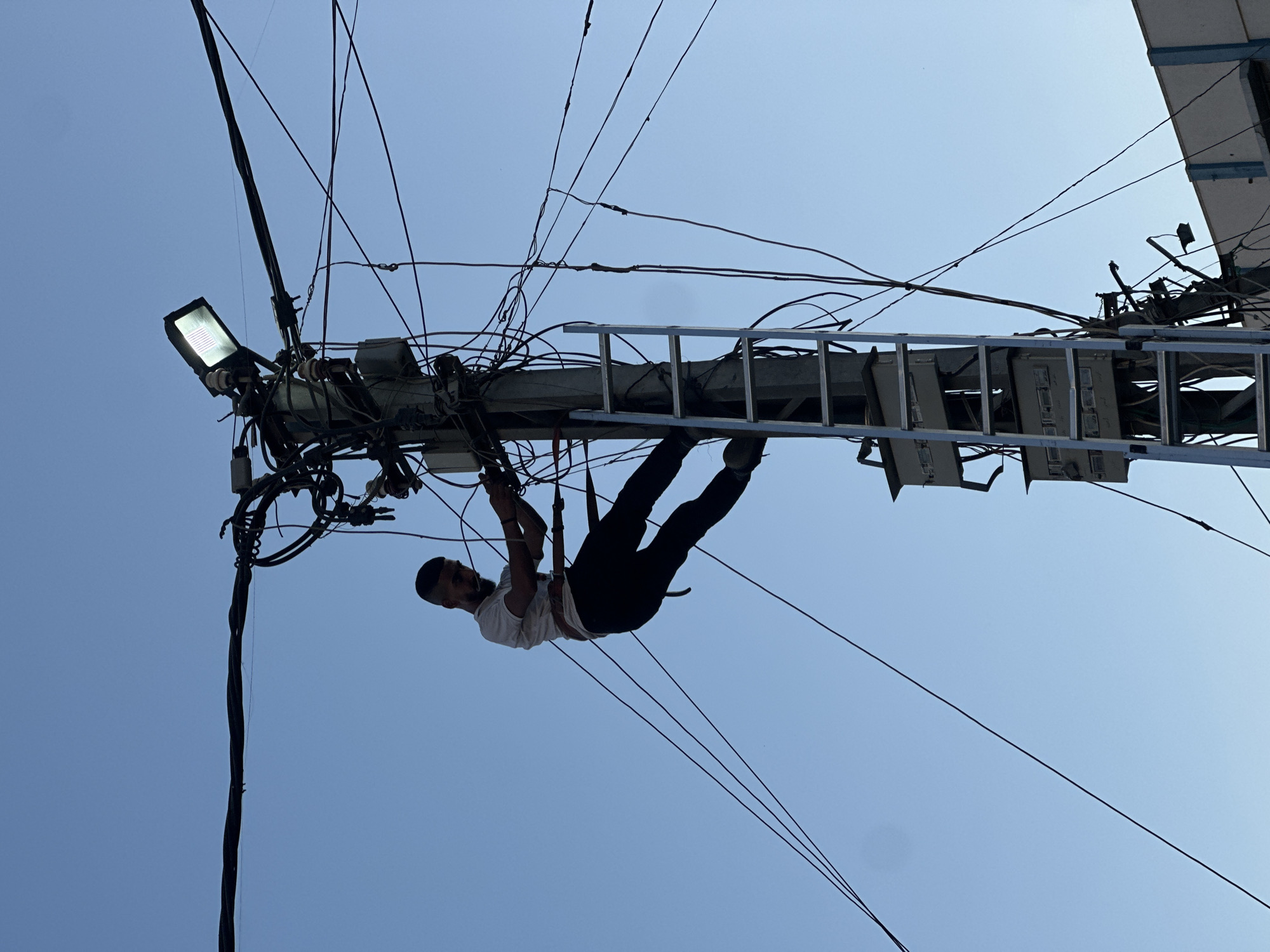 A man is shown climbing a utility pole using a ladder, surrounded by a complex network of wires and electrical components. He is wearing a light-colored t-shirt and dark pants, with a safety belt around his waist. The man appears to be working on the wires, as his hands are engaged with them. A bright floodlight is mounted on top of the pole, illuminating the scene. The sky is a clear, pale blue, indicating daytime. The background shows a building to the right, suggesting an urban or semi-urban environment. The image captures a moment of manual labor in a precarious setting, highlighting the intricate infrastructure of a city.
