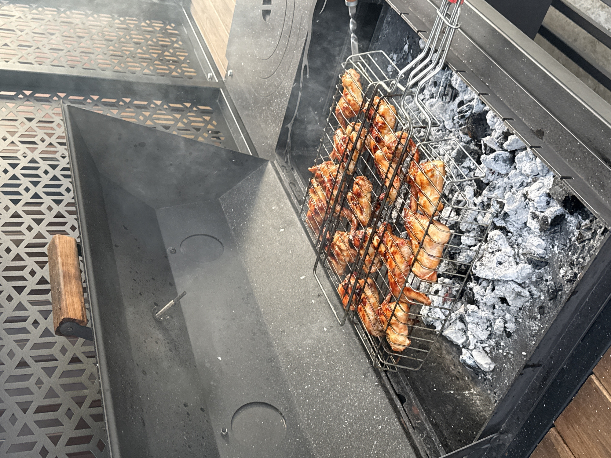 The image depicts a close-up, high-angle shot of chicken wings being grilled in a black, metal barbecue pit. The chicken is held in a wire grill basket placed over glowing embers and ash. The wings appear golden brown and slightly charred, indicating they are close to being fully cooked. Wisps of smoke rise from the grill, suggesting the cooking process is underway.
The barbecue pit has a textured, patterned surface on the left side, and a smooth, dark interior. A wooden handle is attached to the side of the pit on the left. The background shows a portion of the barbecue's lid and what appears to be wooden decking, hinting at an outdoor setting. The lighting suggests it is daytime, likely outdoors, given the presence of the barbecue and natural light. There is no visible text in the image. The overall scene conveys a sense of casual outdoor cooking. The location context provided is Starokorsunskaya, Russia.