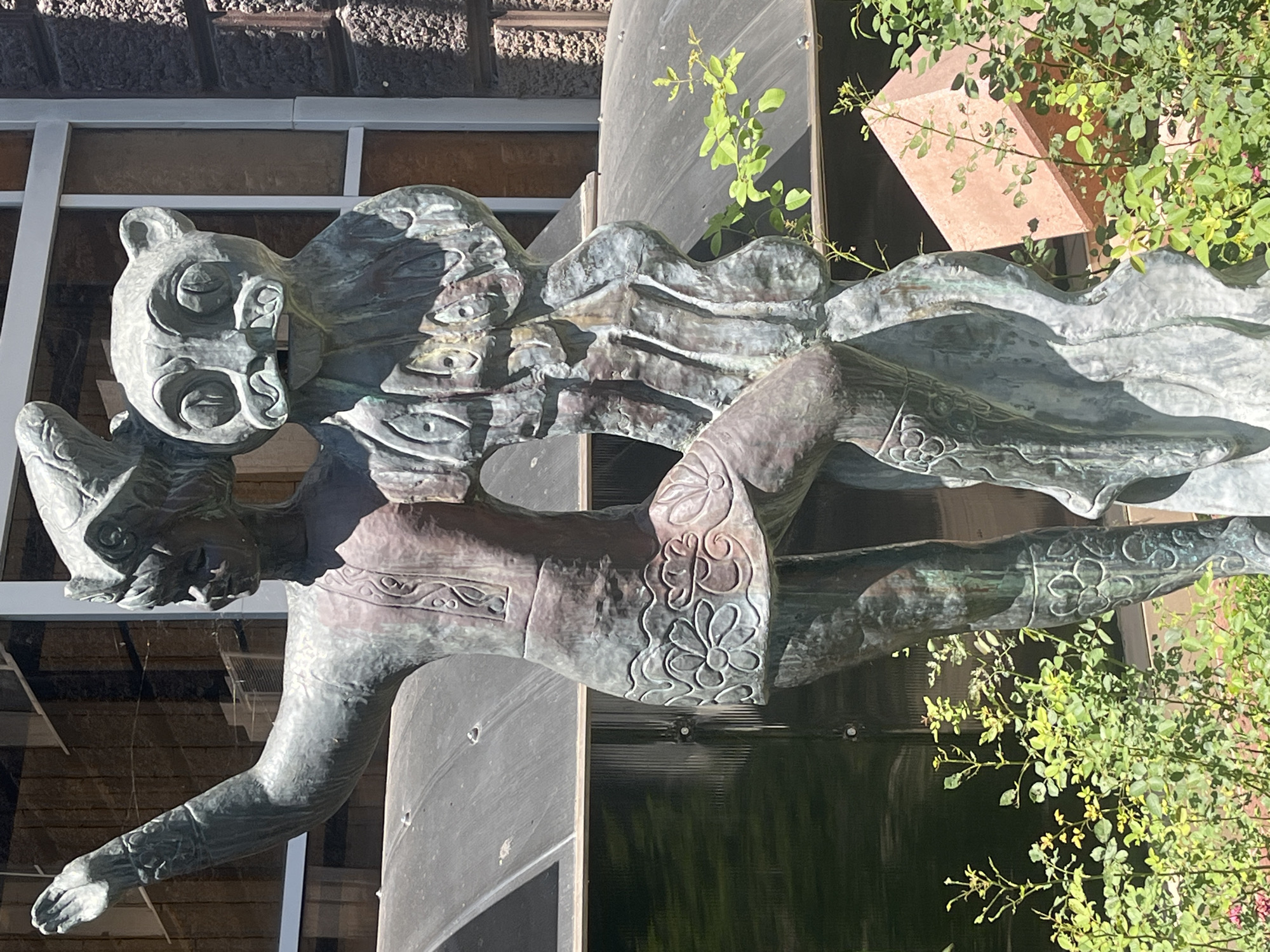 The media file shows a bronze sculpture of two intertwined figures, possibly human or mythical, positioned in an outdoor setting. The sculpture has a weathered patina, indicating it is likely made of aged bronze. One figure has a prominent head with circular features around its eyes and mouth, and a distinct bear-like or feline ear. Its body is lean, with decorative patterns on its chest and arm. The second figure is draped and appears to be in a dynamic pose, its body forming flowing lines that merge with the first. The sculpture is set against a backdrop of a building with large windows and textured walls. Green foliage, likely from a rose bush, is visible around the sculpture, suggesting a garden or landscaped area. A terracotta-colored angular object is also partially visible behind the foliage. The lighting suggests it is daytime, with clear weather. The overall impression is one of an artistic installation in a public space, possibly a park or courtyard, given the presence of greenery and the architectural elements. The location context provided is Krasnodar, Russia. There is no visible text in the image.