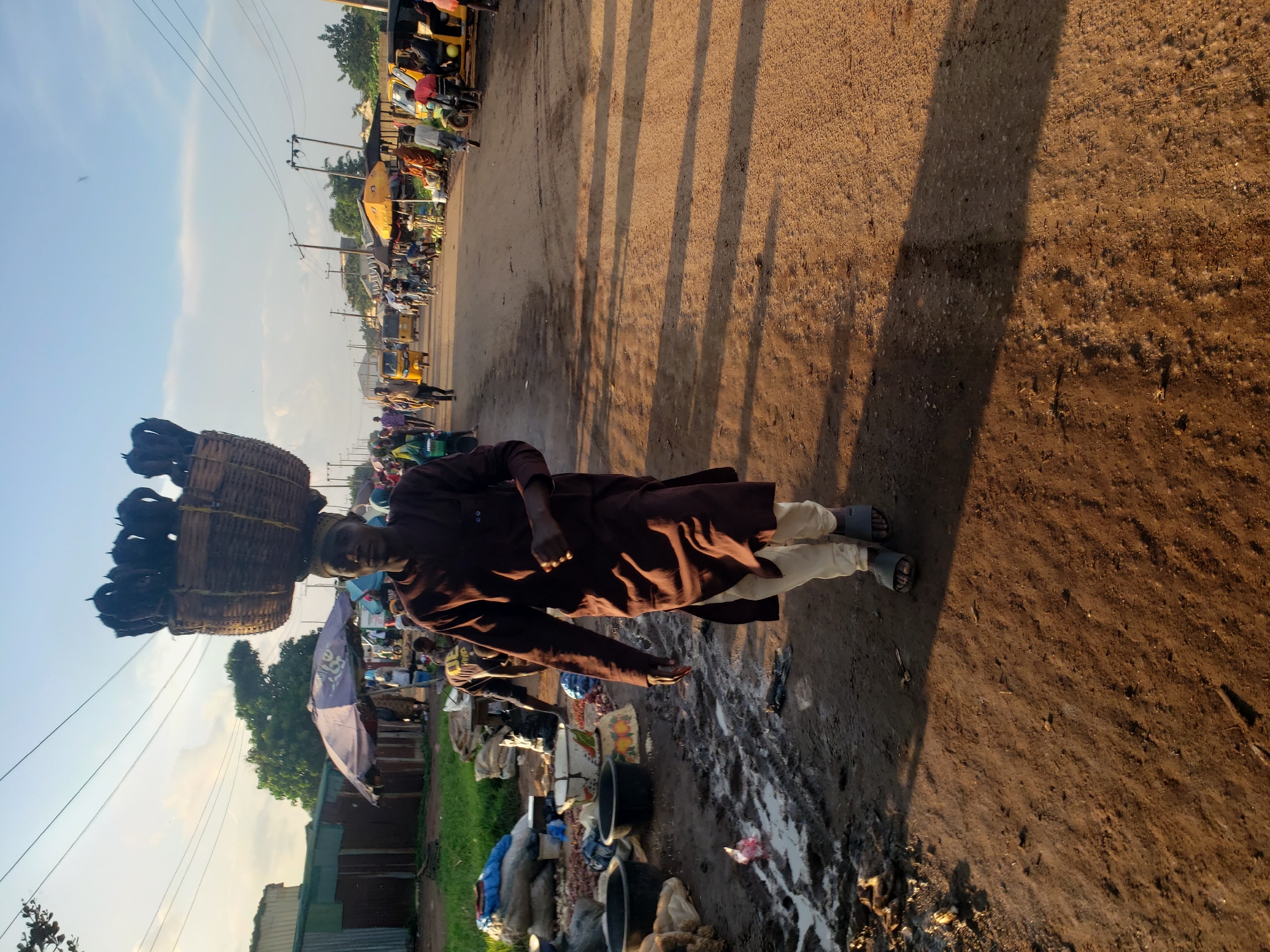 A man walks along a dirt road in Kakuri Gwari, Nigeria, carrying a large woven basket filled with dark objects on his head. He is wearing a long, brown robe and light-colored pants, with dark sandals on his feet. The road is lined with stalls and shops, some covered by umbrellas, and several yellow motorized rickshaws are visible. Bags of produce and other goods are stacked on the left side of the road, and the ground is uneven and muddy in places. The sky is bright and hazy, suggesting it is daytime.