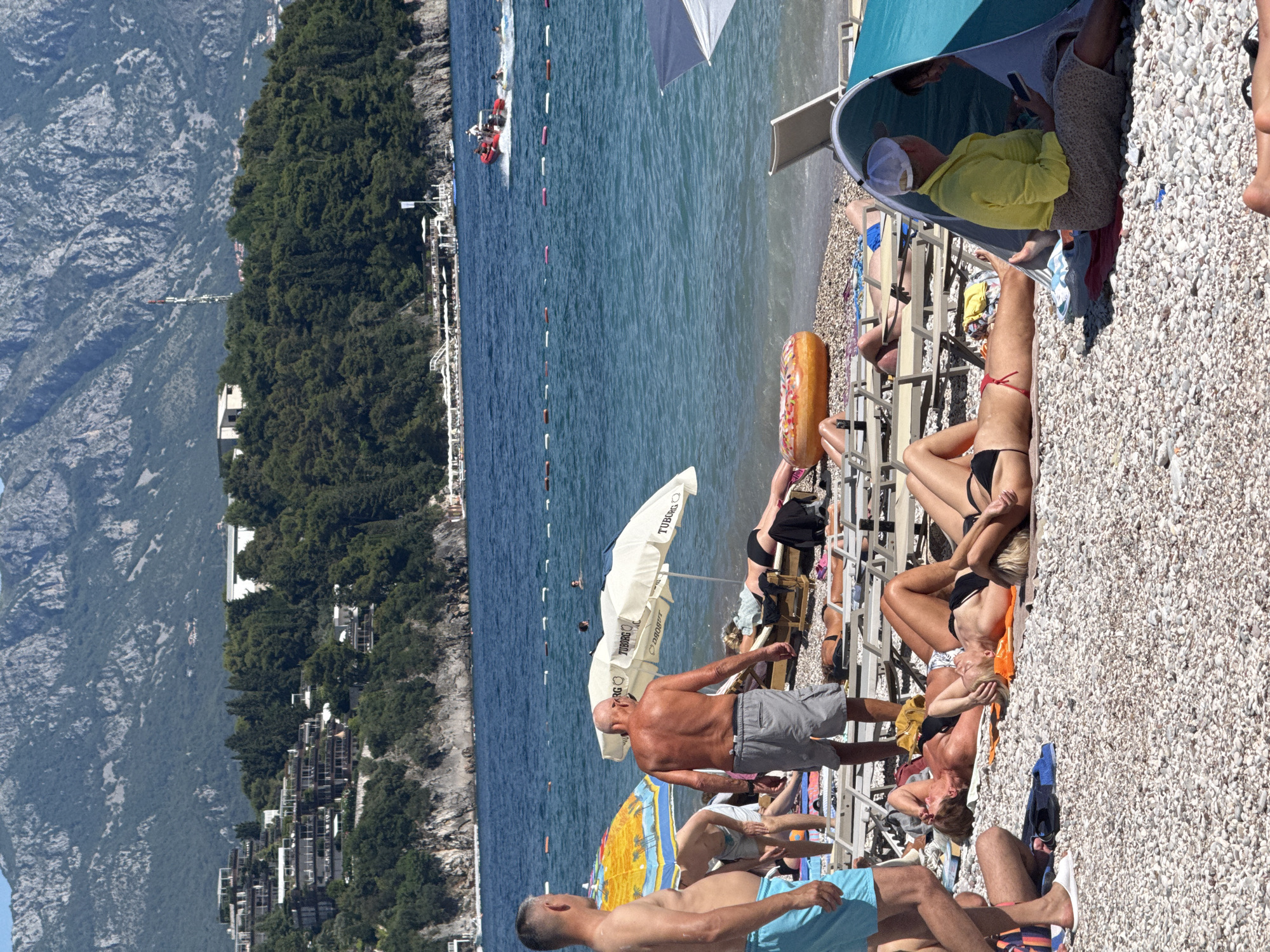 The image depicts a crowded beach scene in Budva, Montenegro. In the foreground, several individuals are sunbathing on lounge chairs and directly on the pebbled beach. A man in blue shorts stands facing away from the camera, while another man in grey shorts is also facing away. Various beach umbrellas provide shade. In the midground, the turquoise sea is visible with a few people swimming. Further out, a red speedboat is cutting through the water, with a person at the helm and another passenger. In the background, the coastline is lined with hotels and residential buildings nestled into a lush, green hillside, which is backed by rugged, grey mountains. The overall atmosphere is one of a busy, sunny day at a popular coastal resort.