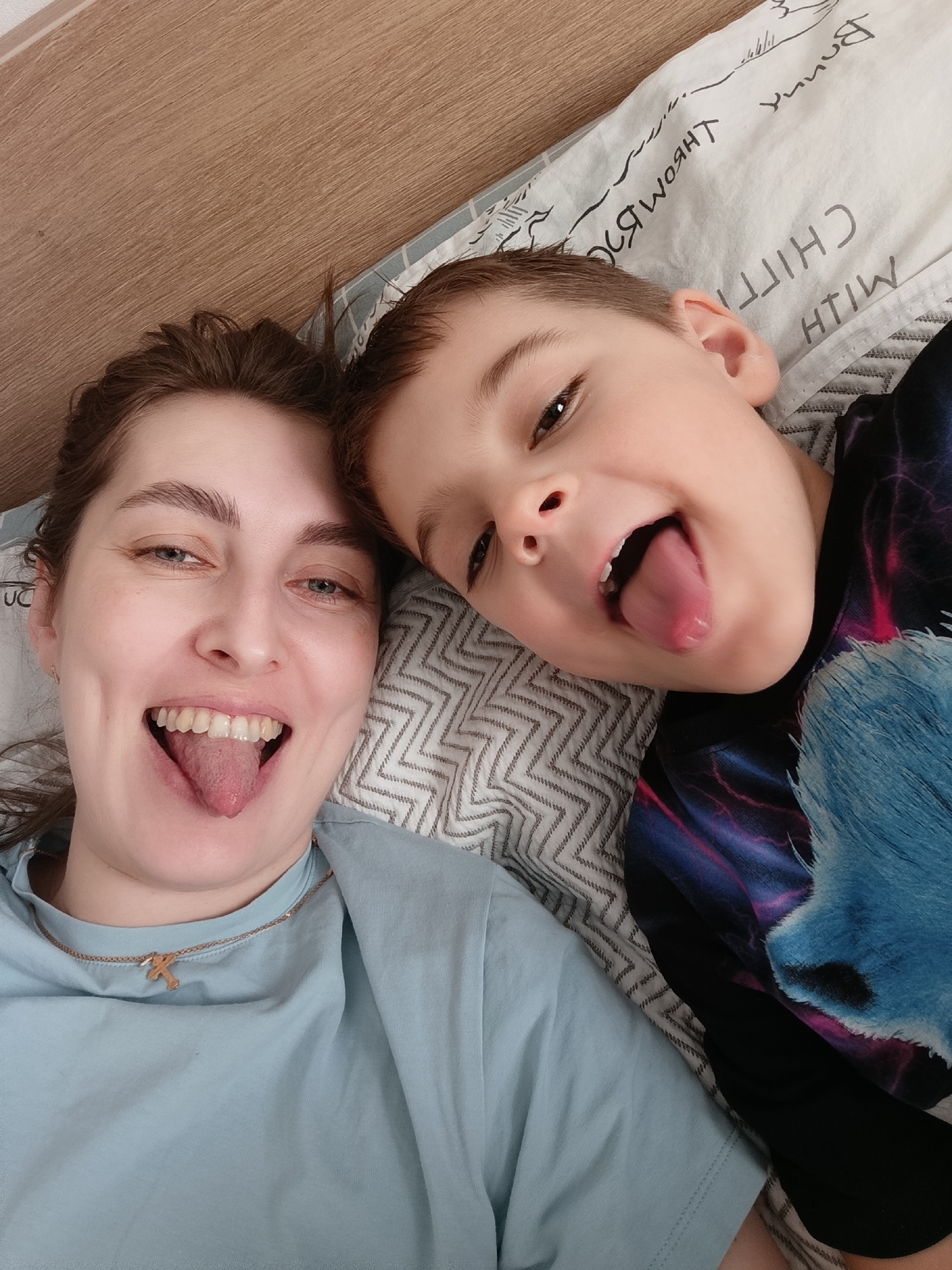 A woman and a child are lying on a bed. They are both sticking their tongues out. The woman is wearing a light blue t-shirt and a gold necklace with a "K" pendant. The child is wearing a shirt with a blue and black design. The bed has a white patterned comforter and is in front of a light brown headboard. It is indoors during the daytime, presumably in Chaikovsky, Russia.