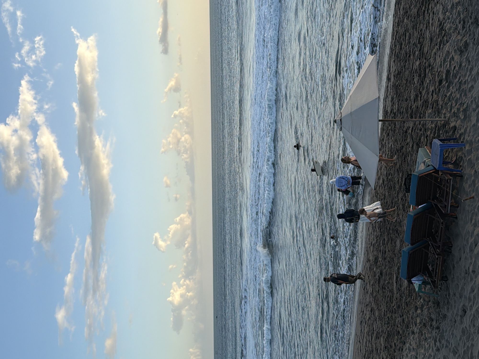 This is a photograph of a beach in North Kuta, Indonesia. The scene captures a section of the coastline with dark sand, calm ocean waters, and a light blue sky dotted with fluffy white clouds. In the foreground, three empty lounge chairs are arranged on the sand, facing the sea. A light-colored umbrella stands beside them, casting a shadow. A small blue table is positioned near the lounge chairs.
Several people are visible on the beach and in the water. On the wet sand near the water's edge, a man in a dark shirt and shorts walks towards the left. Further to the right, a child in shorts walks along the shoreline. A woman in a white skirt and a dark head covering is also present, as is a person in a light blue hooded jacket. In the water, several individuals can be seen. Two surfers are in the distance, one with a surfboard. Another person is swimming closer to the shore. The ocean displays gentle waves with white foam breaking near the beach. In the far distance, small boats are visible on the horizon. The lighting suggests it may be late afternoon or early evening.