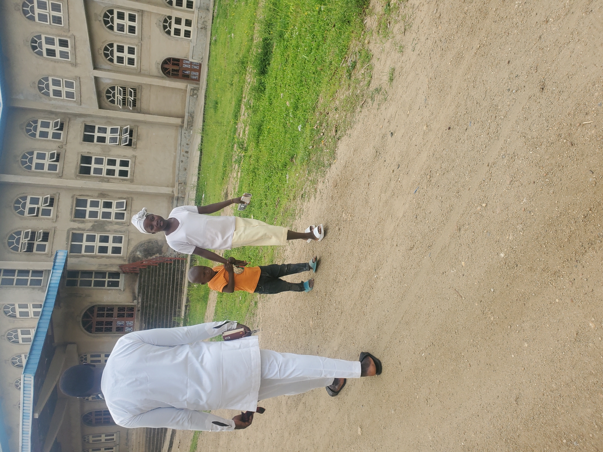 In Kabwir, Nigeria, an adult male in a white kaftan and pants walks away from the camera, holding a dark object in his right hand. To his right, a woman in a white headscarf and t-shirt walks towards the camera, holding the hand of a young boy. The boy, wearing an orange t-shirt and dark pants, walks alongside the woman, also towards the camera. They are walking on a dirt path in front of a light-colored, multi-story building with arched windows. Green grass borders the path.
