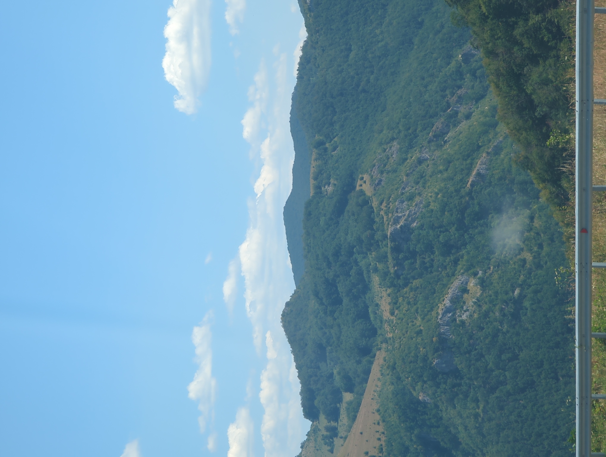 The image depicts a panoramic view of lush green mountains under a bright blue sky dotted with scattered white clouds. The terrain is varied, with steep slopes covered in dense forestation, interspersed with rocky outcrops and some areas of sparse vegetation on drier slopes. In the foreground, a metal guardrail runs horizontally across the bottom of the frame, suggesting the viewpoint is from a road or elevated vantage point. The scene is peaceful and natural, characteristic of a mountainous landscape. There are no people or man-made structures visible in the mountainous area. The lighting indicates it is daytime, likely a sunny afternoon. Given the context provided, this scene is likely located in the Vratsa region of Bulgaria, known for its dramatic mountain scenery.