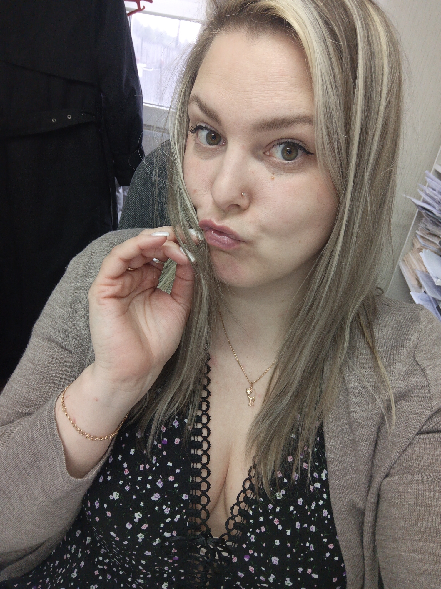 A selfie depicts a woman with fair skin, hazel eyes, and blonde hair with light highlights. She has a nose piercing and is wearing eyeliner. Her expression is playful, with pursed lips. She wears a gold necklace featuring a cat pendant, a gold bracelet, a black floral dress with lace trim, and a beige cardigan. Her fingernails are long and painted white.
Behind her, there is a dark-colored garment hanging and a stack of papers. The location is Noviy, Russia.