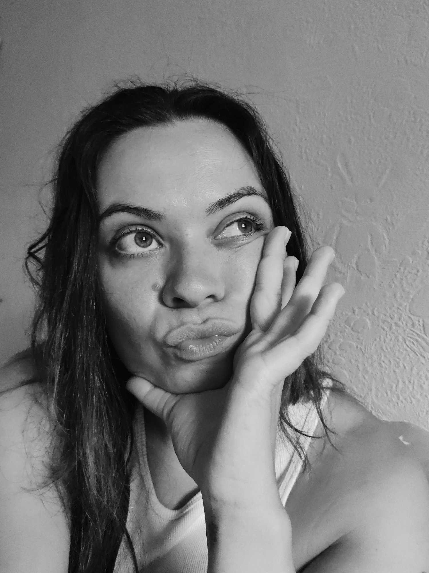 A black and white, close-up, medium shot photo shows a woman in her late 20s or early 30s. She is facing right, with her head tilted up and her hand on her chin. Her lips are pursed, and her eyes are looking upwards and to the right. She has dark, medium-length hair that is slightly messy and falls around her face and shoulders. She is wearing a light-colored tank top. The background is a textured wall. There is no information available about the location of the image in Chaikovsky, Russia.