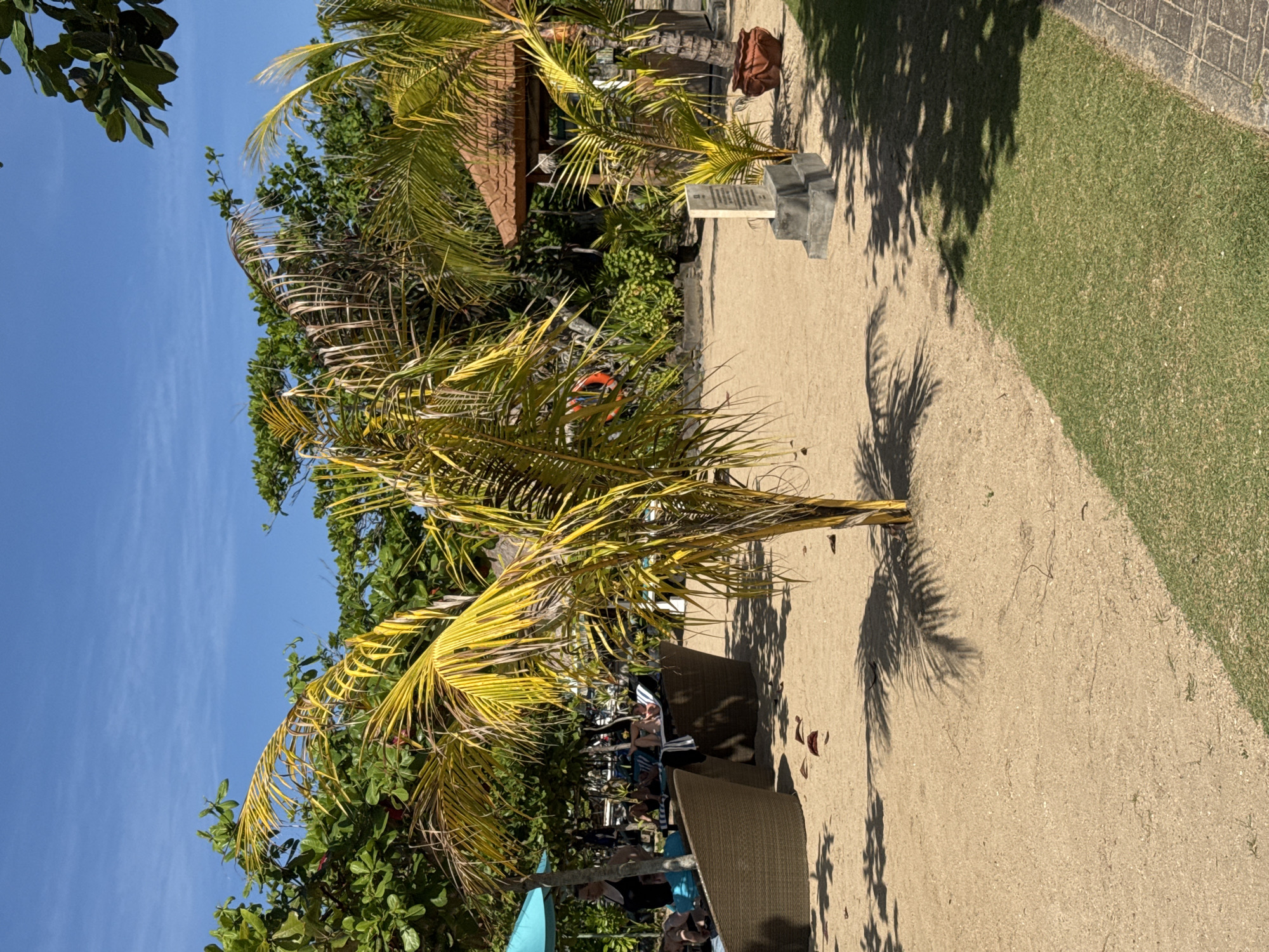 The image captures a sunlit scene on a sandy path, possibly near a beach in Kuta Selatan, Indonesia. The sky is blue, visible behind foliage. Palm trees, one with yellowing fronds, dominate the foreground. People are seated in chairs under umbrellas, suggesting a leisure or relaxation area. To the right of the frame, a patch of green grass borders the sandy path. Further in the background, a small structure with a tiled roof is visible amidst denser vegetation. A stone marker sits at the edge of the path.