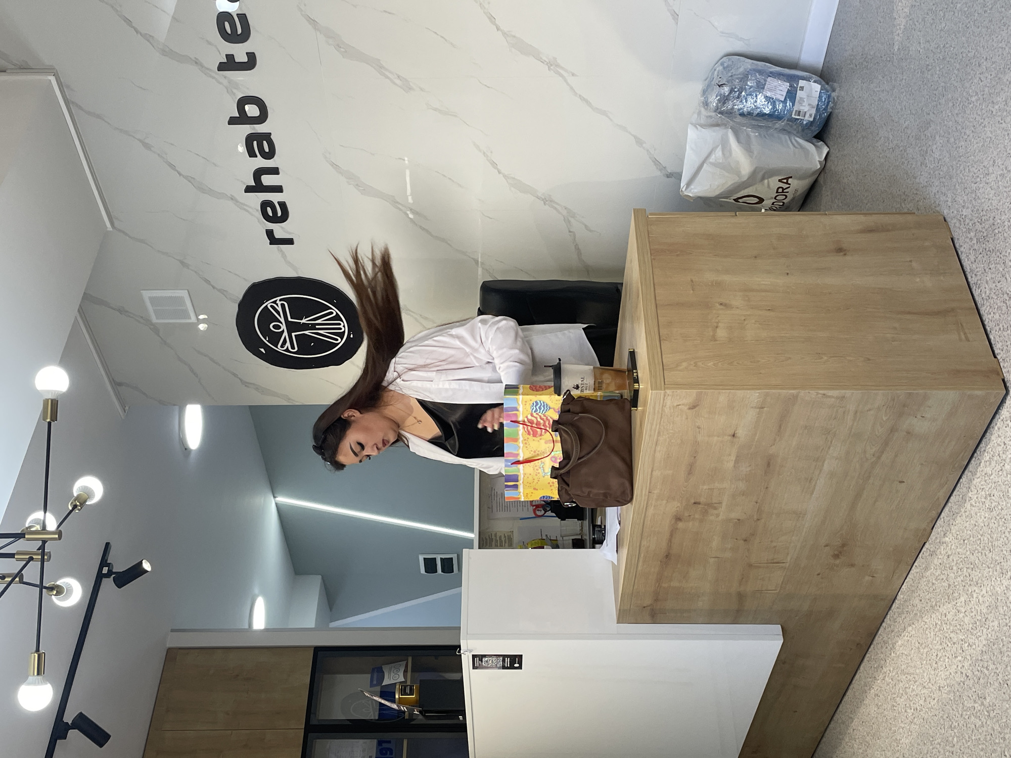A woman with long dark hair is standing behind a wooden reception desk. Her hair is being blown to the side. She is wearing a white shirt over a black top and a necklace. On the desk, there is a brown purse, a colorful gift bag, and a cup of iced coffee. A logo with a figure in a circle is visible on the wall behind her, along with the text "rehab te". There are also two white bags and a blue plastic-wrapped item on the floor to the right of the desk. The environment appears to be a modern clinic or office space with marble-patterned walls and a light-colored floor. The location is identified as Almaty, Kazakhstan.
