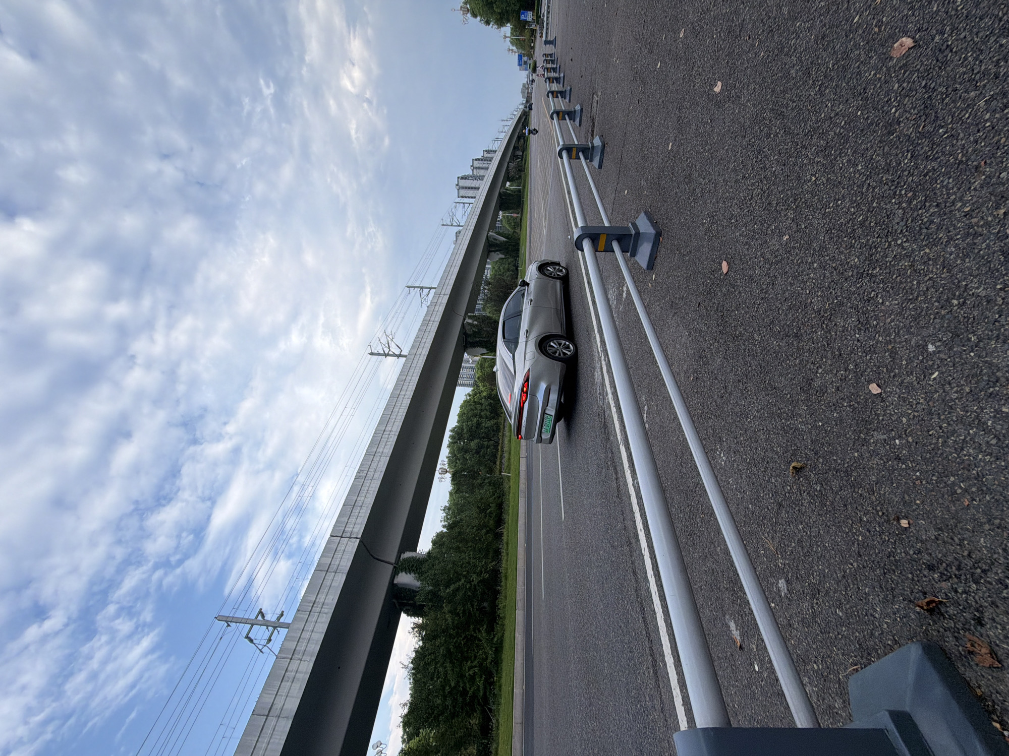 A silver sedan is traveling on a multi-lane asphalt road in Jiangjin, China, under an elevated railway track. The car is in the right lane of the road, moving away from the camera. The road is marked with white lines. In the foreground, a metal guardrail with gray posts separates the road from a paved area. In the background, lush green trees line the side of the road, and beyond them, tall buildings are visible. The sky is overcast with a mix of clouds and blue sky. The scene appears to be in an urban or suburban area.