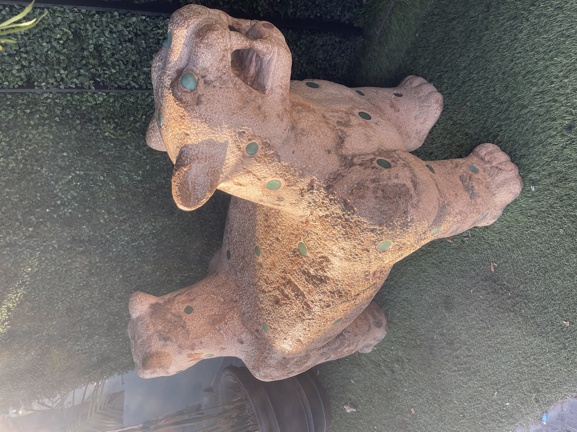 A stone or concrete statue of a jaguar lies on artificial turf. The statue is mottled brown and has green circular inlays representing the jaguar's spots. The statue's head faces the top left, and its four paws are splayed out. The background includes a green artificial hedge. Bottom left corner shows the wheel of a stroller. The location is in Playa del Carmen, Mexico.