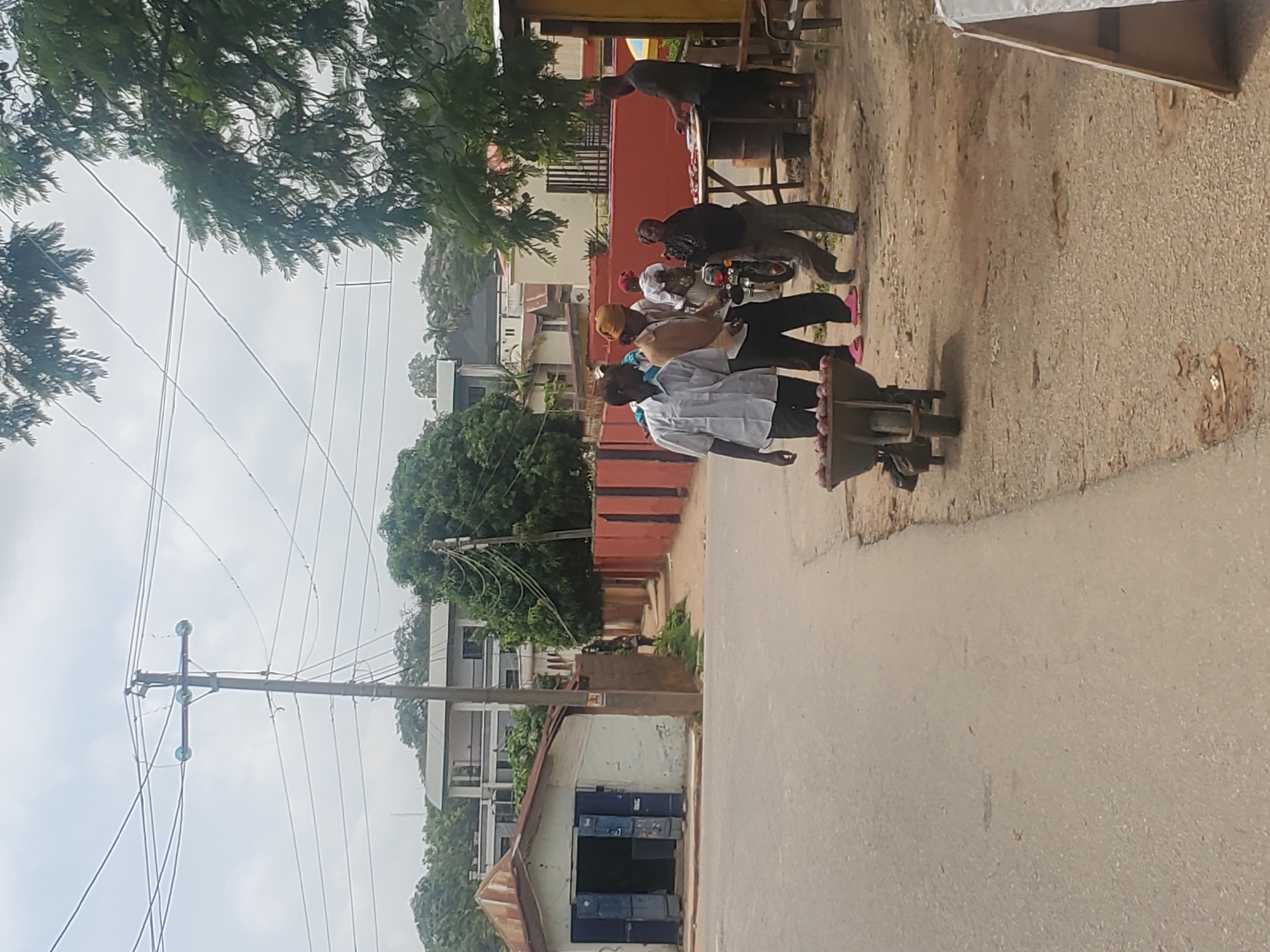 A group of people are gathered near roadside stalls, with a motorcycle parked among them. The scene is set along a dirt road next to a paved surface, suggesting a commercial area or market. In the background, there are buildings, trees, and utility poles with overhead wires, indicating an urban or semi-urban environment. The people appear to be engaged in some form of exchange or interaction, with some individuals standing and others seated or on the motorcycle. One person is behind a stall displaying items, possibly food or goods. Another person is wearing a white shirt and dark pants, and another is in a dark shirt and dark pants. Pink flip-flops are visible on one person's feet. The overall atmosphere seems casual, with no strong indications of a specific event or urgent activity. The time of day appears to be daytime, judging by the lighting, and the weather seems to be overcast or hazy. No visible text is present in the image.
