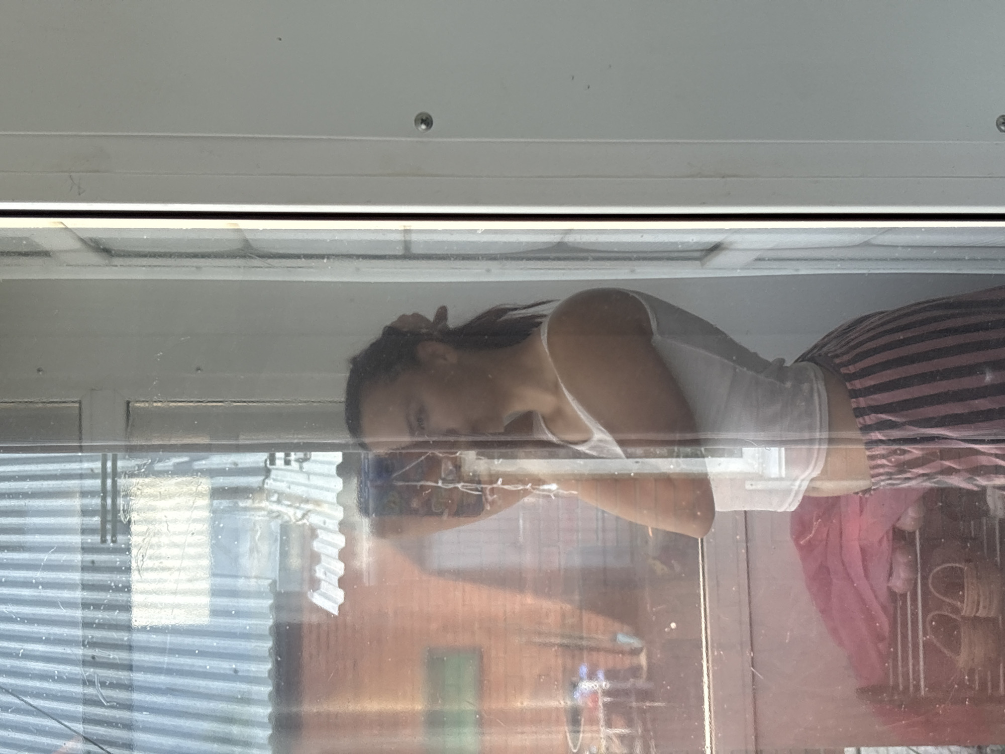A person is captured in a reflection on a transparent surface, possibly glass or plastic. They are wearing a white tank top and striped pants. The person's head is tilted back, and their hand is resting on their head. Their gaze appears to be directed towards a phone they are holding, suggesting they are taking a selfie.
The background is somewhat obscured by the reflection and the transparent material, but it appears to be an indoor setting with blinds or a similar window covering. Outside, there are glimpses of buildings and possibly other structures. The lighting suggests it is daytime.
The image is characterized by the distorted and layered visuals due to the reflection and the transparent surface. The colors are muted, with the prominent pink and black stripes of the pants and the white of the tank top standing out. The overall mood is casual and personal, typical of a self-portrait. There is no visible text in the image.