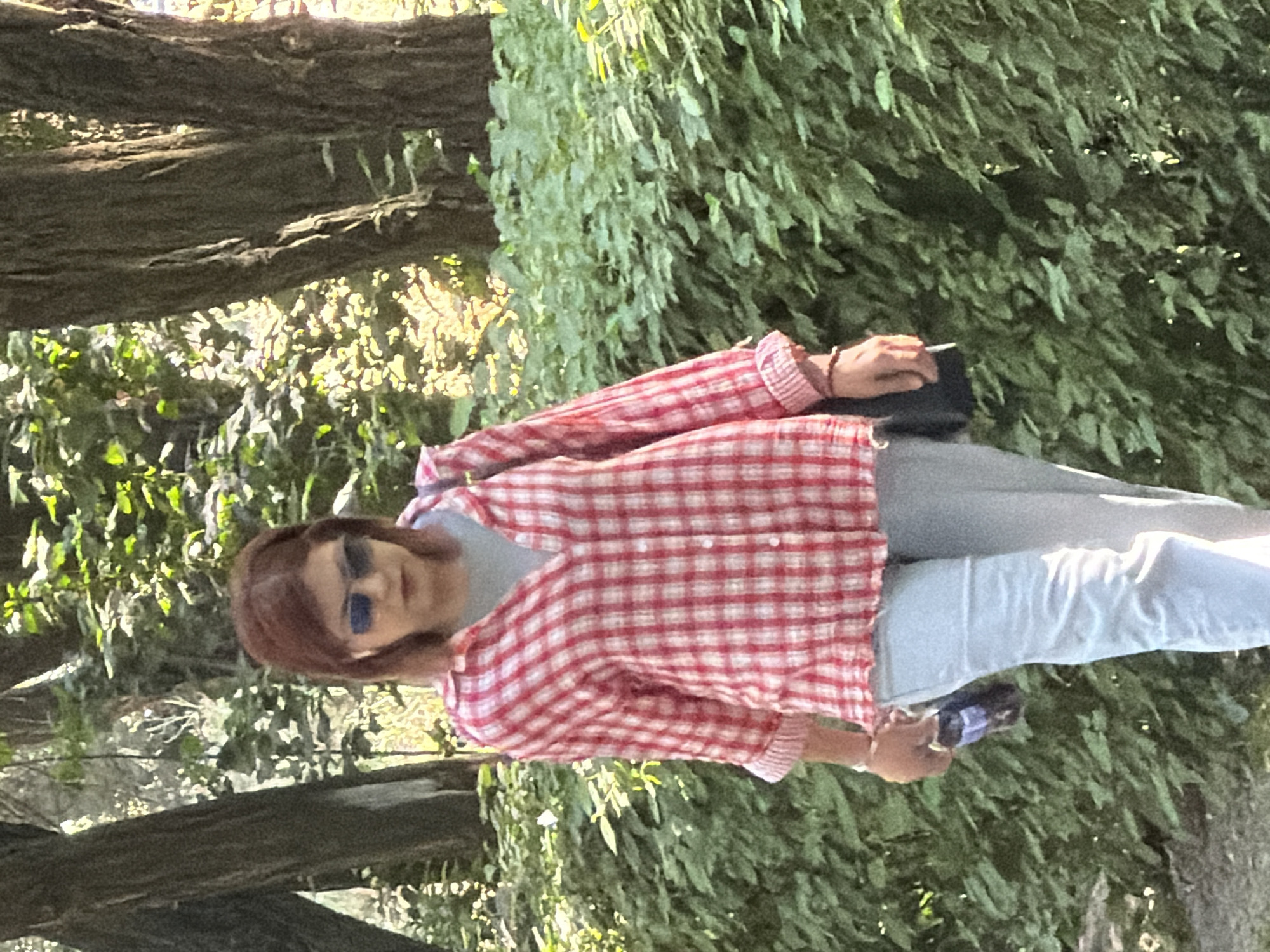 A middle-aged woman with reddish-brown hair, wearing sunglasses, a red and white plaid shirt, and light blue pants, walks through a park. She is holding a dark-colored object in her right hand and has a dark bag slung over her right shoulder. The background consists of trees and lush green foliage, illuminated by sunlight.