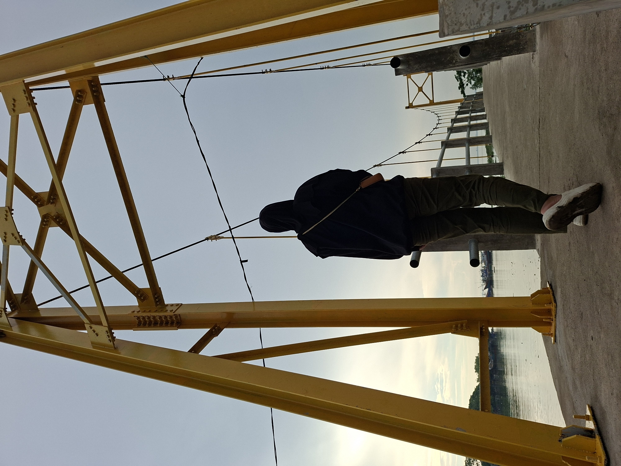 A person wearing a black hijab and a dark blue hoodie walks away from the camera on a concrete bridge. They are wearing olive green pants and white sneakers. The person is walking towards a suspension bridge with yellow metal support structures. In the background, a river is visible with several boats docked on the far side, and a cloudy sky above. The scene appears to be an outdoor walkway in Lhokseumawe, Indonesia, possibly part of a bridge or pier infrastructure near the waterfront.