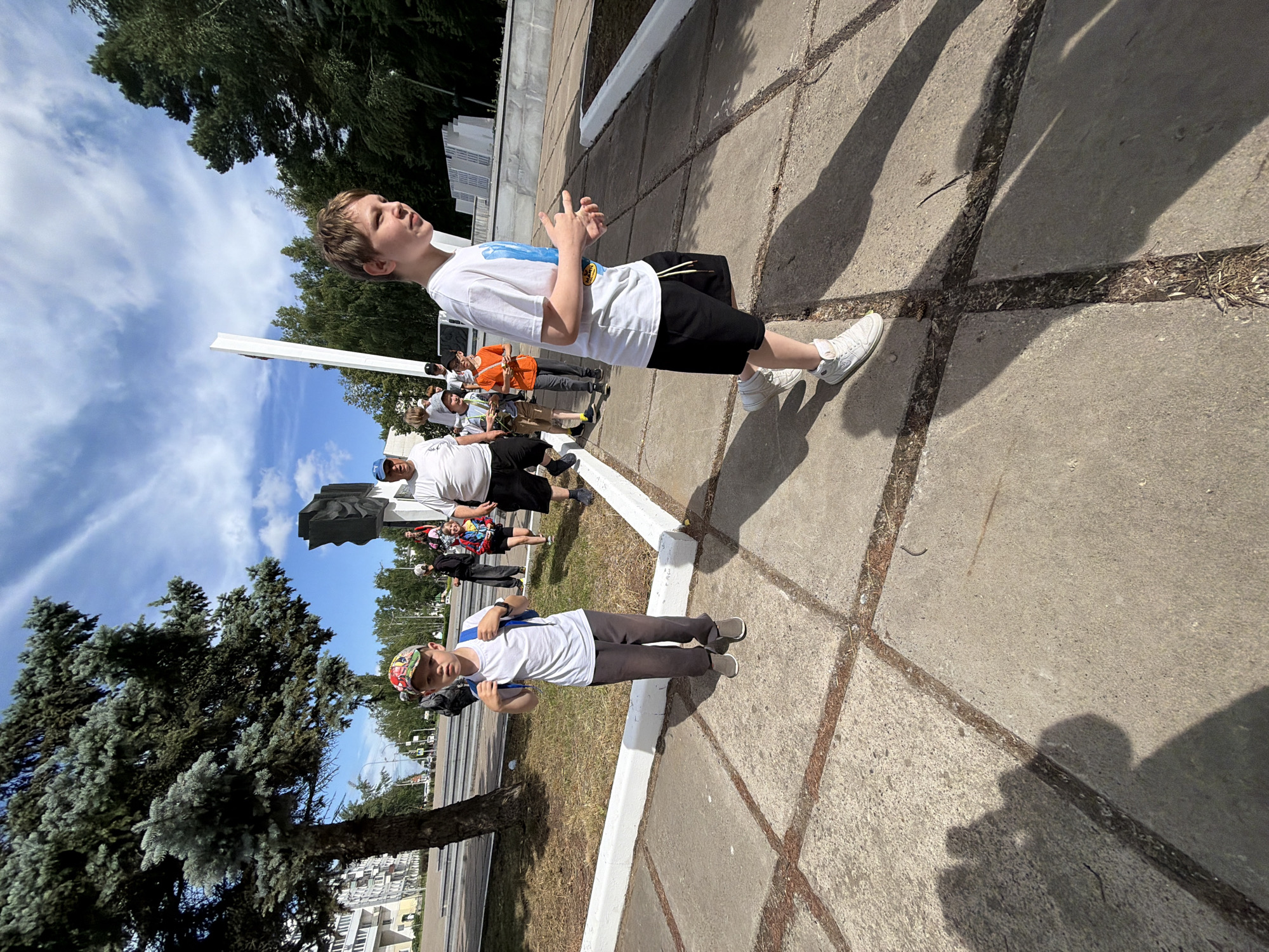 In Chaikovsky, Russia, a group of people, including several children, walk on a paved area near a large tree and a monument. The children in the foreground are wearing backpacks and casual clothing. One boy in a white t-shirt and black shorts, wearing white high-top sneakers, is facing away from the camera and appears to be gesturing with his hands. Another boy in a white t-shirt, grey pants, and a colorful cap is looking towards the camera. A man in a white t-shirt and a blue cap is visible in the background, along with other individuals. The environment features a clear sky with scattered clouds, trees, and buildings in the distance. The ground is paved with square concrete slabs.