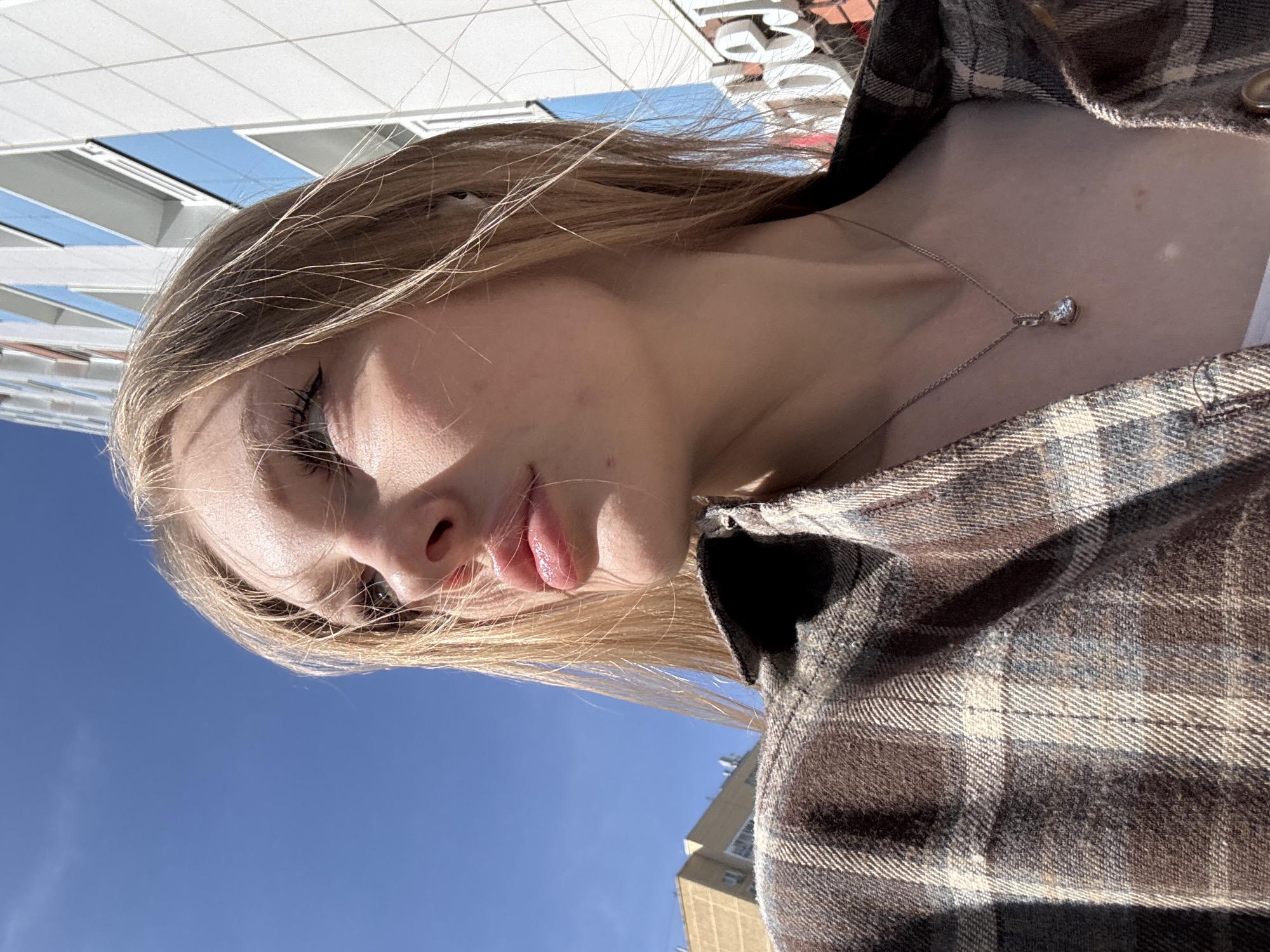A close-up, tilted, low-angle shot shows a young woman outdoors. She has blonde hair and is wearing a plaid shirt and a silver necklace. The background includes a clear blue sky, a building with a white facade, and a partial view of a structure in the distance. The exact location in Perm, Russia is not deducible from the image.