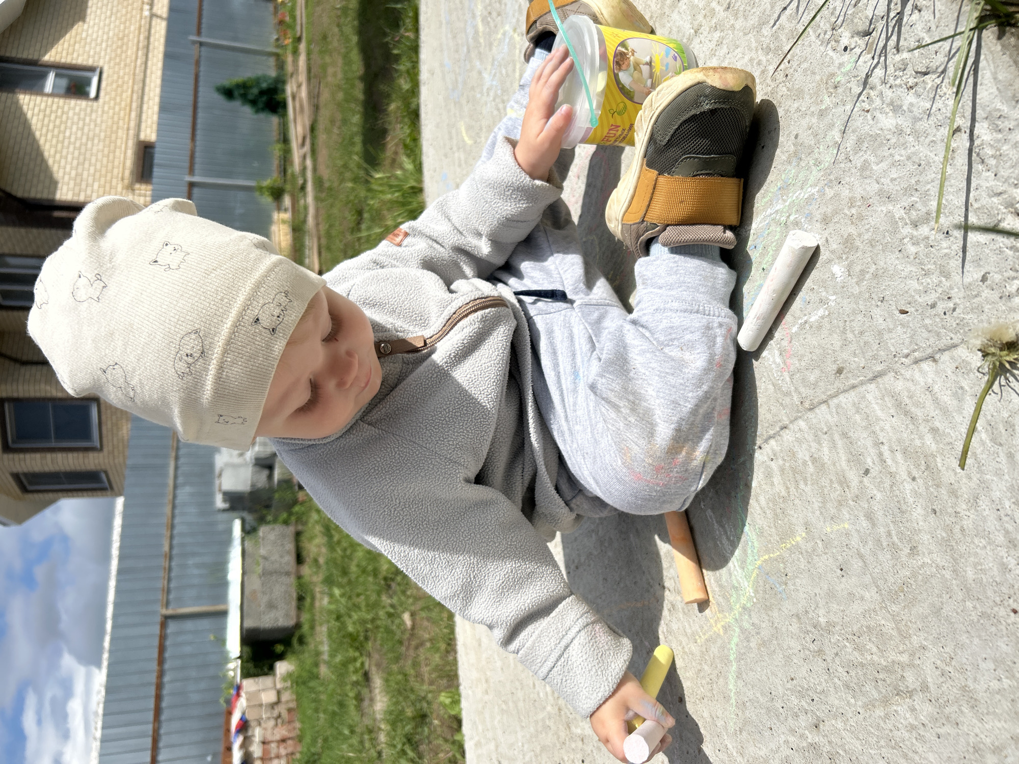 A young child, wearing a light-colored beanie with a repetitive animal pattern, a grey fleece jacket, and grey sweatpants, is sitting on a concrete surface outdoors. The child is holding a yellow piece of chalk in their right hand and appears to be drawing on the concrete, with other pieces of chalk, including an orange one and a white one, visible nearby. Colorful chalk marks, predominantly green and yellow, are scattered on the concrete around the child. A yellow plastic container with a blue lid, which may contain more chalk or art supplies, is positioned to the child's right. The child is also wearing brown and grey sneakers. In the background, a brick house with windows is visible, along with a metal fence and green grass. The sky is partly cloudy. The scene suggests an outdoor art activity by a child in a residential area. The presence of chalk art on a concrete surface is characteristic of areas where children play, such as courtyards or sidewalks. The backdrop of a house and fence indicates a domestic or suburban setting.