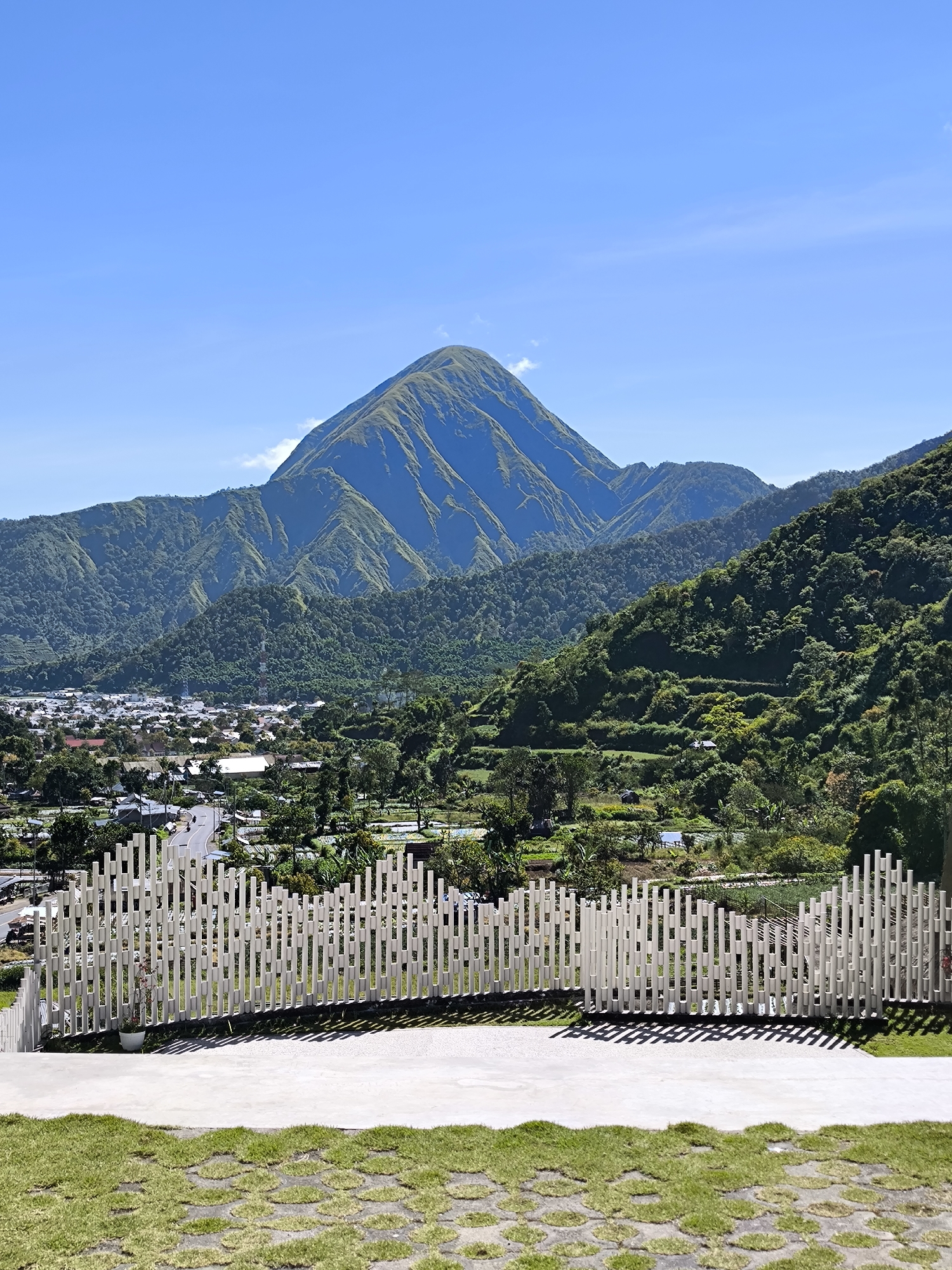 The image shows a mountain landscape in Sembalun Bumbung, Indonesia, viewed from an elevated platform with a decorative white fence. The mountain, with a green, grassy peak, dominates the background. Several lower, forested hills lead towards the mountain. In the mid-ground, there's a view of a town with buildings and roads. In the foreground, a concrete platform is bordered by a textured grass surface. The sky is clear and blue.