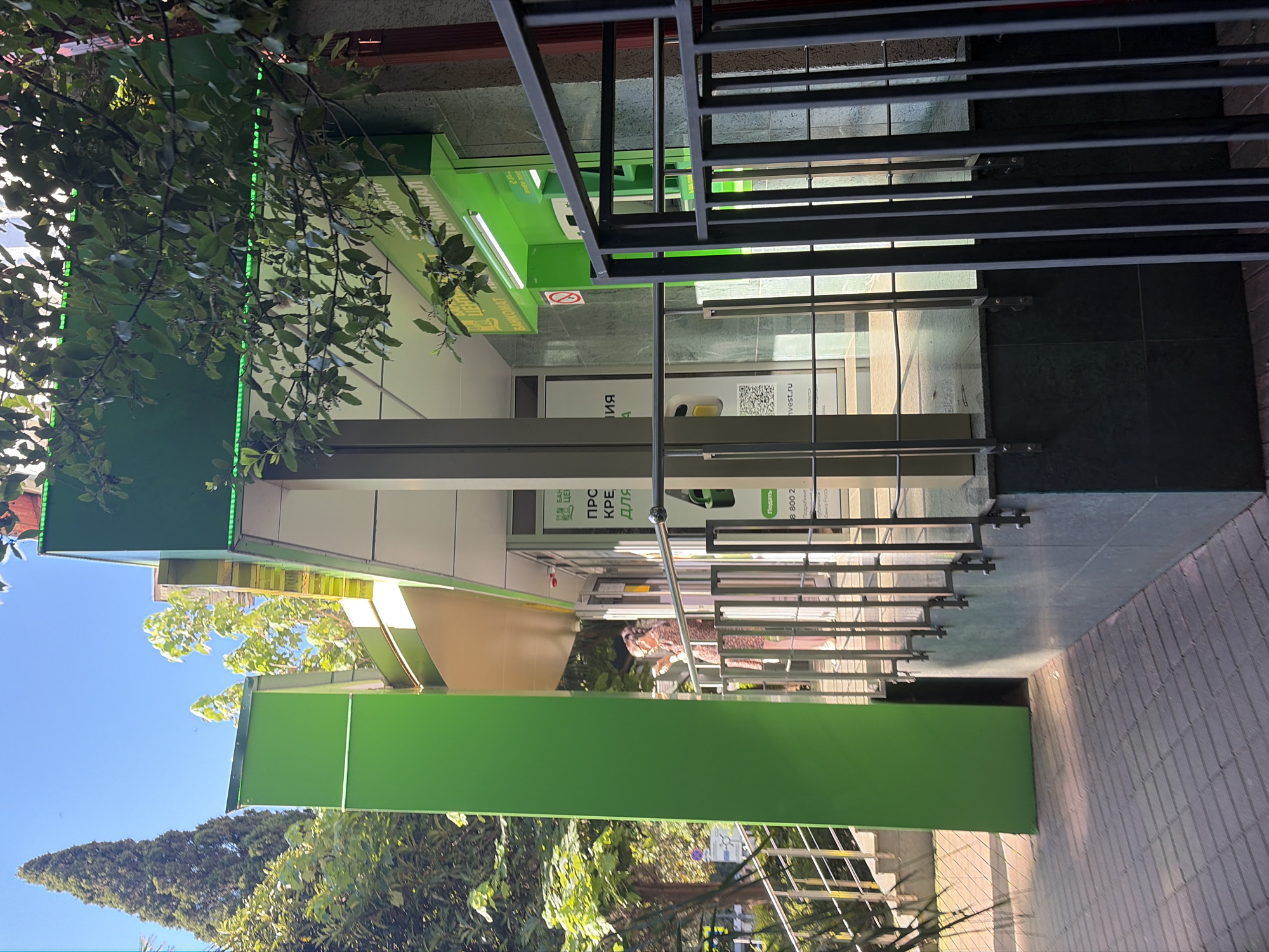 The image depicts an outdoor, daytime view of a modern entrance, likely to a bank or an ATM lobby, distinguished by its prominent green branding elements. The scene is set under a clear blue sky, with abundant green foliage from trees and bushes partially overhanging the building from the left and appearing in the foreground and background.
The entrance features large glass doors and panels, revealing a brightly lit interior with fluorescent lighting. An ATM machine, predominantly green, is visible through the glass. The building's facade incorporates significant green structural components and signage. A dark metal railing or fence with horizontal bars is prominent on the right side, leading towards the entrance, alongside shiny silver handrails. The ground in the foreground is paved with light-colored rectangular bricks.
Several textual elements are visible. On a green sign, partially obscured by leaves, "БАНКОМАТ" (ATM) is discernible, along with part of a phone number starting with "8 800". Inside, through the glass, a sign reads "ПРОДАЖА КРЕ..." (Sale of credit/loans or similar financial products), accompanied by a QR code and the text "invest.ru". A red circular "no smoking" symbol is also clearly visible near the ATM. No individuals are present in the image. The overall impression is that of a clean, accessible financial service point, likely a Sberbank location given the color scheme and typical branding in Russia.