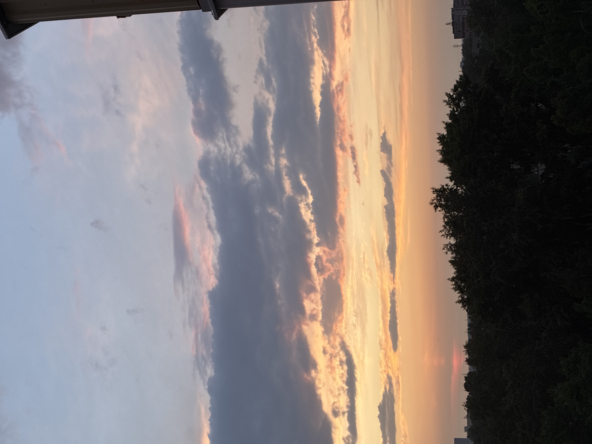 A sunset view shows a silhouetted line of dark green trees against a sky filled with orange and pink clouds. The sky transitions from a bright orange near the horizon to lighter hues of pink, purple, and blue higher up. A tall, narrow, light-colored structure is visible on the right side of the image, partially obscuring the sky. Distant buildings and power lines can be seen beyond the treeline on the left and right sides of the frame.