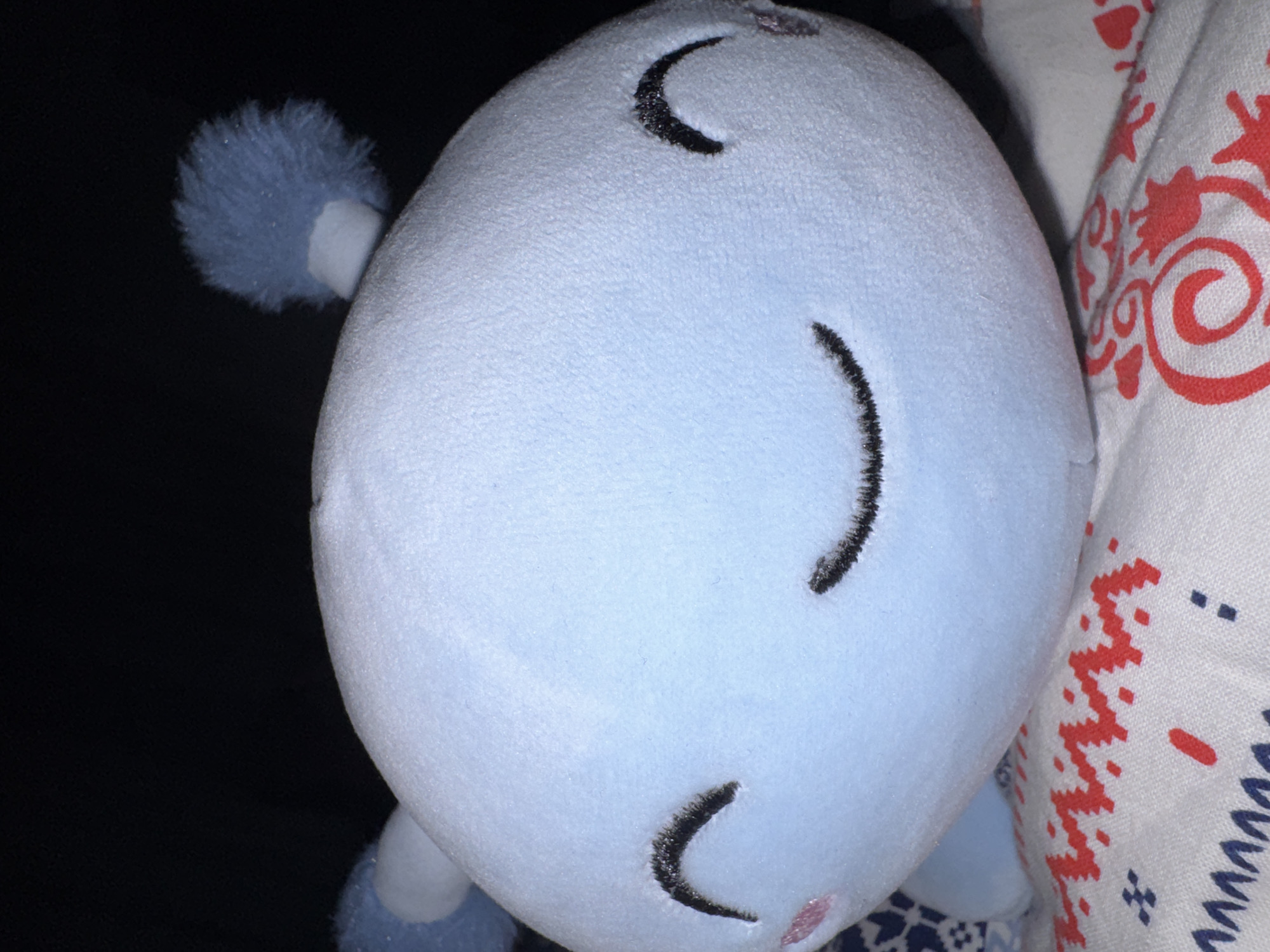 A close-up, slightly tilted image captures a light blue, oval-shaped plush toy appearing to be asleep. The toy's face is rotated ninety degrees counter-clockwise, featuring three embroidered black crescent shapes that depict closed eyes and a tranquil, sleeping mouth. A subtle pink marking, possibly a nose or blush, is visible below the lowest crescent.
On the left side of the plush toy, a small, fluffy, light blue pom-pom, which could be an ear or a tail, is visible against a dark background. The plush material exhibits a soft, short-pile texture. To the right of the toy, a white fabric with a detailed pattern of red and blue designs is present. These patterns include swirls, floral motifs, geometric shapes, and zigzags, suggesting a blanket, pillowcase, or similar textile.
The scene appears to be indoors, under low light conditions, possibly indicating evening or nighttime. The general mood is one of peace and rest. There are no people visible, nor any specific activity taking place other than the implied sleep of the toy. No text is visible in the image.