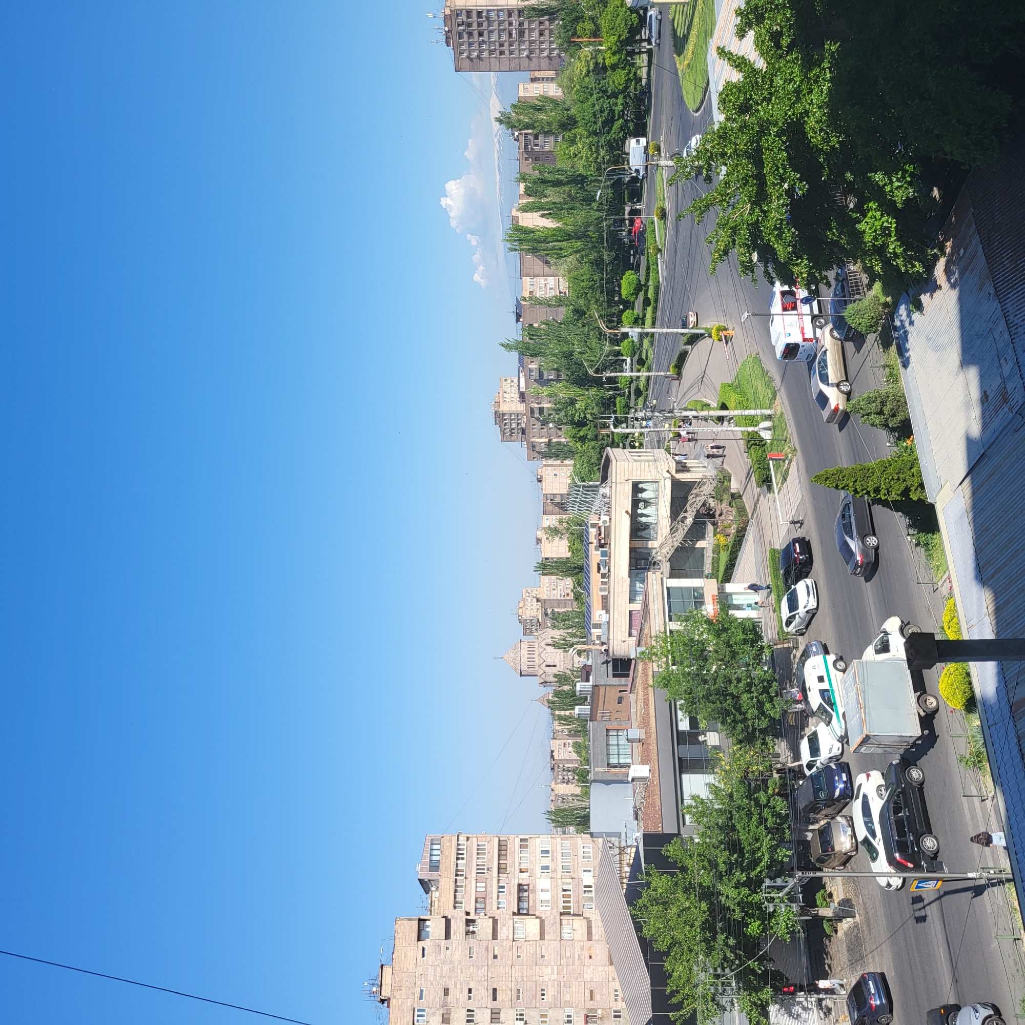 The media file presents an elevated, wide-angle view of a bustling urban street scene in Yerevan, Armenia, on a bright, sunny day. The image is taken from a building overlooking a multi-lane road that curves through the city.
In the foreground and mid-ground, a busy street is visible with numerous vehicles, including cars, vans, and a large white truck with a gray cargo bed. An ambulance, white with red markings, is also seen on the road. Traffic lights and streetlights line the thoroughfare, which is bordered by manicured green spaces and tall, leafy trees. Several people can be seen walking on the sidewalks and along the street. On the right, the corrugated metal roof or balcony edge of the viewpoint is partially visible.
Beyond the immediate street level, multi-story buildings of varying architectural styles dominate the landscape. Many appear to be concrete residential or commercial structures, possibly from the Soviet era, interspersed with more modern facades. Towards the left of the image, a distinctive religious structure with traditional Armenian architectural elements, resembling a cathedral (possibly Saint Gregory the Illuminator Cathedral), stands out amidst the other buildings.
The background is dominated by a vast, clear blue sky with only a few wisps of white clouds. In the far distance, towering mountains are visible, their peaks partially obscured by a light haze or clouds, suggesting the iconic presence of Mount Ararat, a significant landmark in the region. The overall impression is one of a vibrant city enjoying clear weather. There is a blue and yellow pedestrian crossing sign visible on the right side of the road.