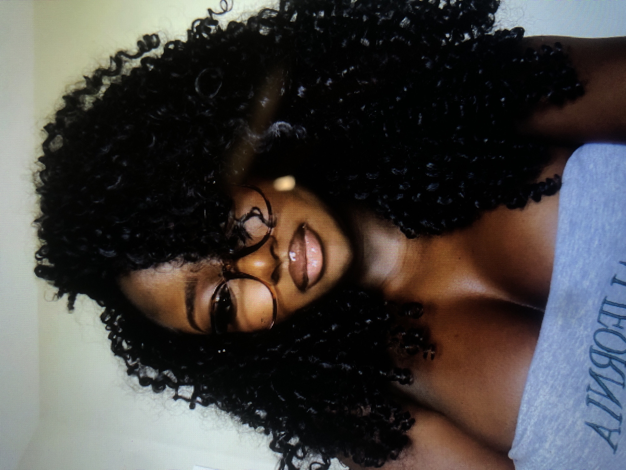 This media file features a close-up, tilted shot of a woman with dark, curly hair, wearing round-framed glasses and a light-colored garment with visible text. The woman's face is the primary focus, with her lips slightly parted and her gaze directed away from the camera, suggesting a moment of reflection or engagement with something outside the frame. The lighting appears to be soft and diffused, creating highlights on her skin and lips. The background is a plain, light-colored surface, offering no specific environmental cues. The visible text on her garment appears to be in a serif font, oriented vertically. The overall impression is intimate and personal, with a focus on the subject's features and style. The context provided for Barakin Lerawi, Nigeria, offers a geographical location but no specific visual elements within the image directly confirm this setting.