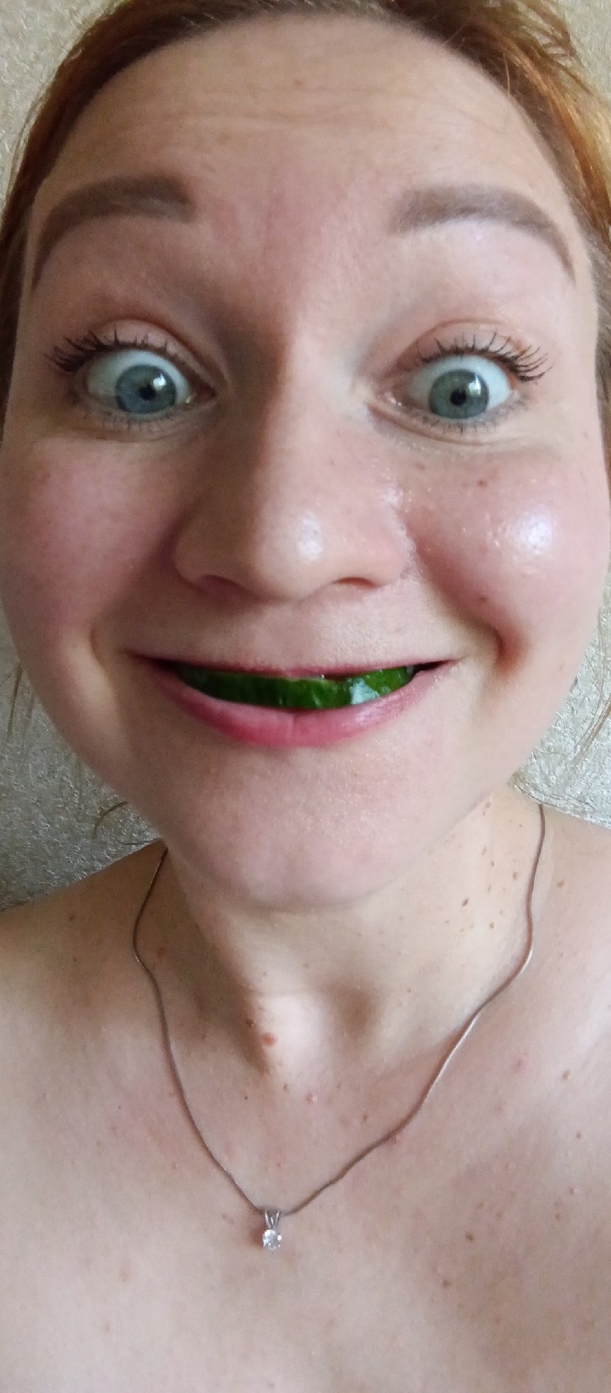 The image shows a close-up of a woman's face, with a slice of cucumber held between her lips. She has fair skin, blue eyes, and reddish hair. Her eyes are wide, and she is smiling, revealing the cucumber. A silver necklace with a pendant is visible around her neck. The background is a plain, light-colored wall. The location is Unknown, Unknown.
