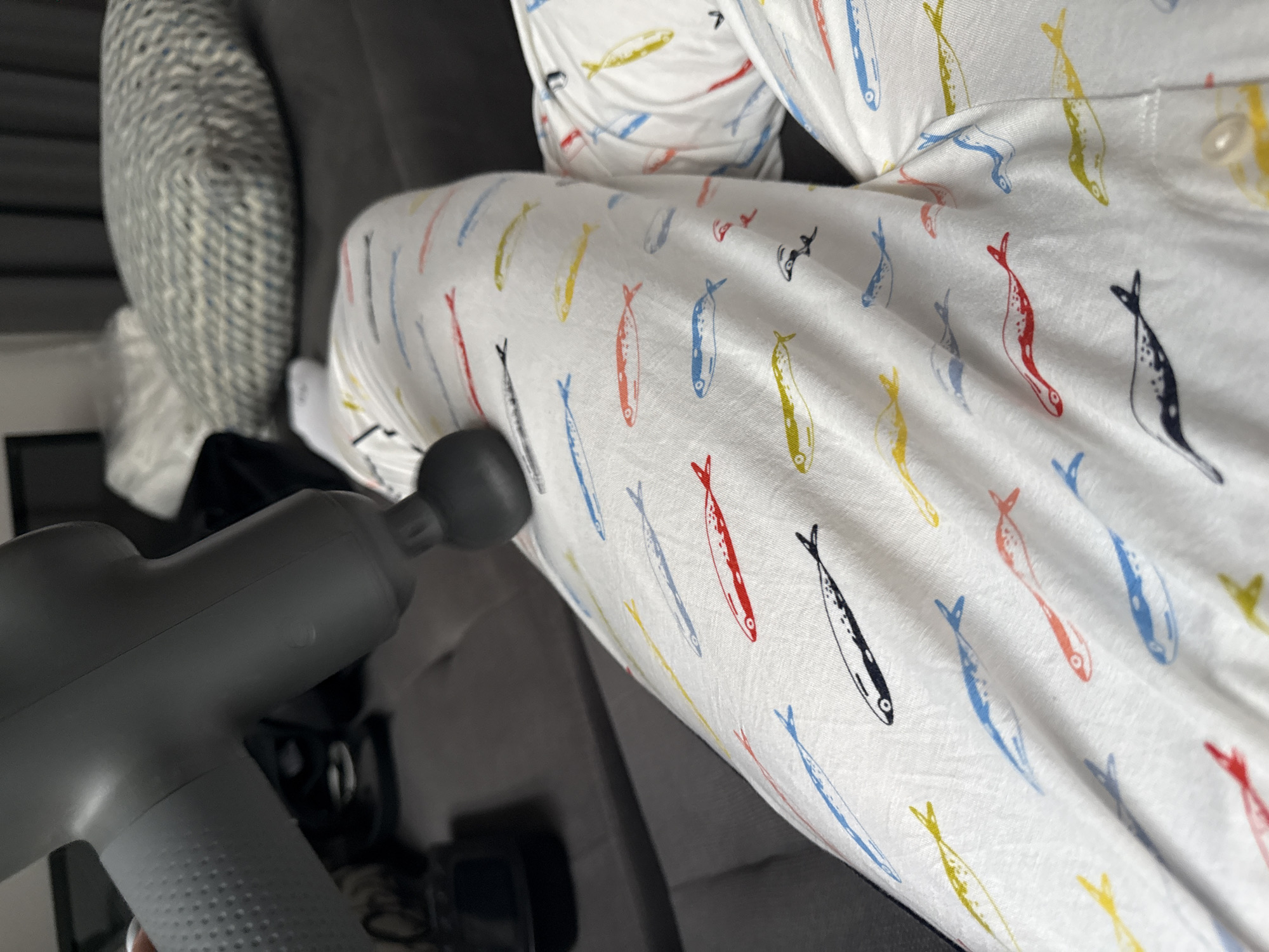A gray massage gun is being used on a person's leg. The person is wearing white pajamas with a pattern of colorful fish. The leg is resting on a gray sofa next to a textured gray pillow. The location is not evident in the image.