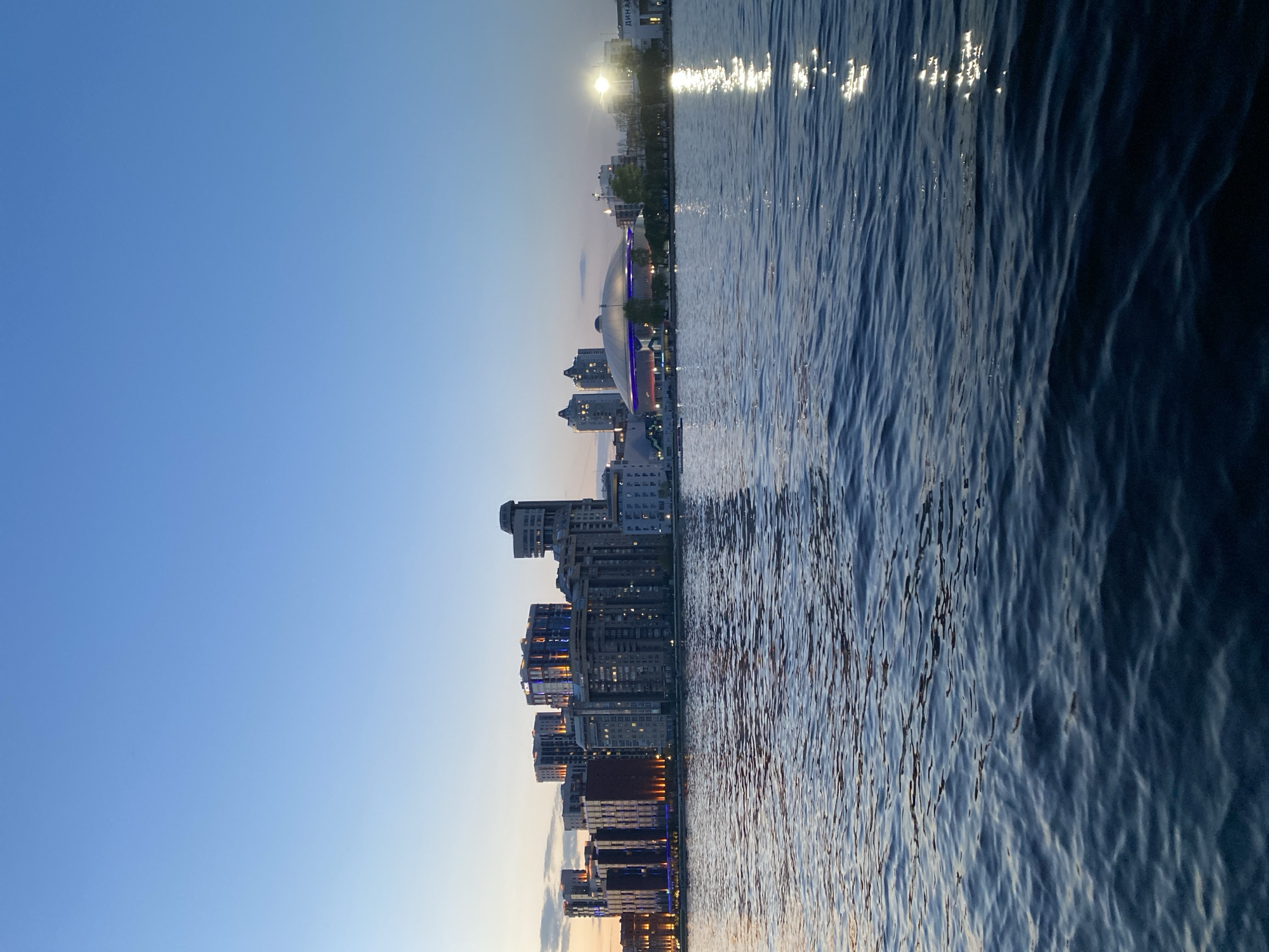 The image shows a waterfront cityscape at dusk. On the left side of the image are multiple buildings, possibly apartments or offices, of various heights. The lights are visible inside some of the apartments and the exterior lights illuminate parts of the building. The sky is a gradient of blue, with darker blue transitioning to a lighter blue. The right side of the image is water, reflecting both the city buildings and the sky, with ripples indicating movement. There is an area where the sun reflects off the water. The location is the Iset River in the city of Yekaterinburg, Russia.