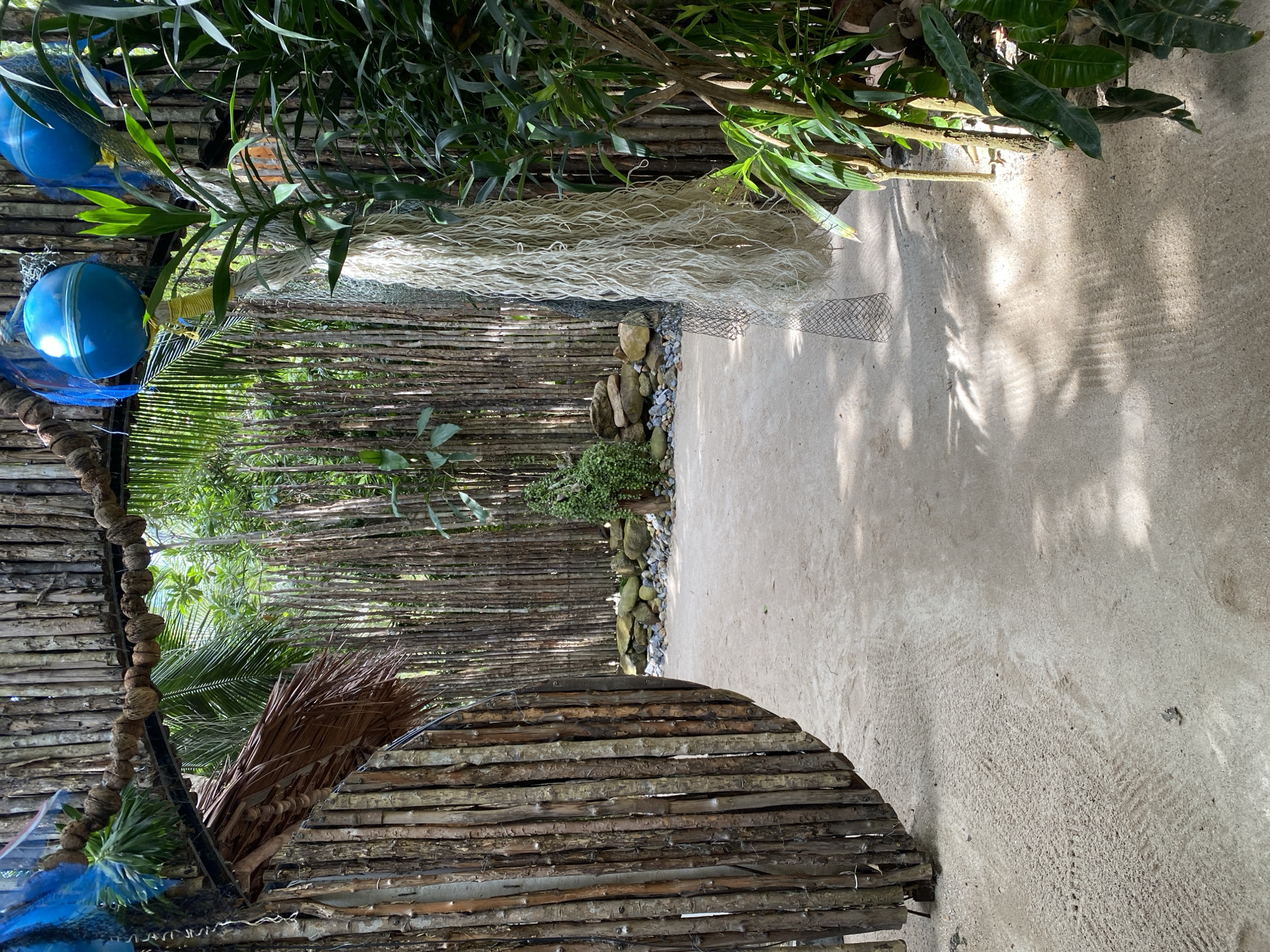 The image depicts an outdoor scene with a focus on natural materials and tropical vegetation. In the foreground, a curved structure made of horizontal wooden logs forms a barrier or enclosure. To the left, two bright blue buoys are visible, partially obscured by netting. Beyond this, a densely packed wall of vertical bamboo or similar slender logs creates a naturalistic backdrop. Lush green foliage, including palm fronds and other tropical leaves, dominates the upper portion of the image, casting dappled shadows on the ground.
A sandy or concrete pathway runs along the right side of the image, leading into the distance. This path is textured with faint patterns, possibly from previous foot traffic or the material itself. Along the pathway, a scattering of stones and a small, meticulously arranged pile of larger rocks create a decorative element. A long, thick strand of pale, rope-like material, possibly dried plant fiber, lies draped over the wooden structure and extends across the scene. A fine mesh net is also visible in the middle ground.
The overall impression is one of a rustic, naturalistic setting, possibly an outdoor seating area, pathway, or decorative installation within a tropical environment. The bright sunlight and strong shadows suggest it is daytime, likely with clear weather. The location context of Koh Phangan, Thailand, reinforces the tropical ambiance. No people or text are visible in the image.