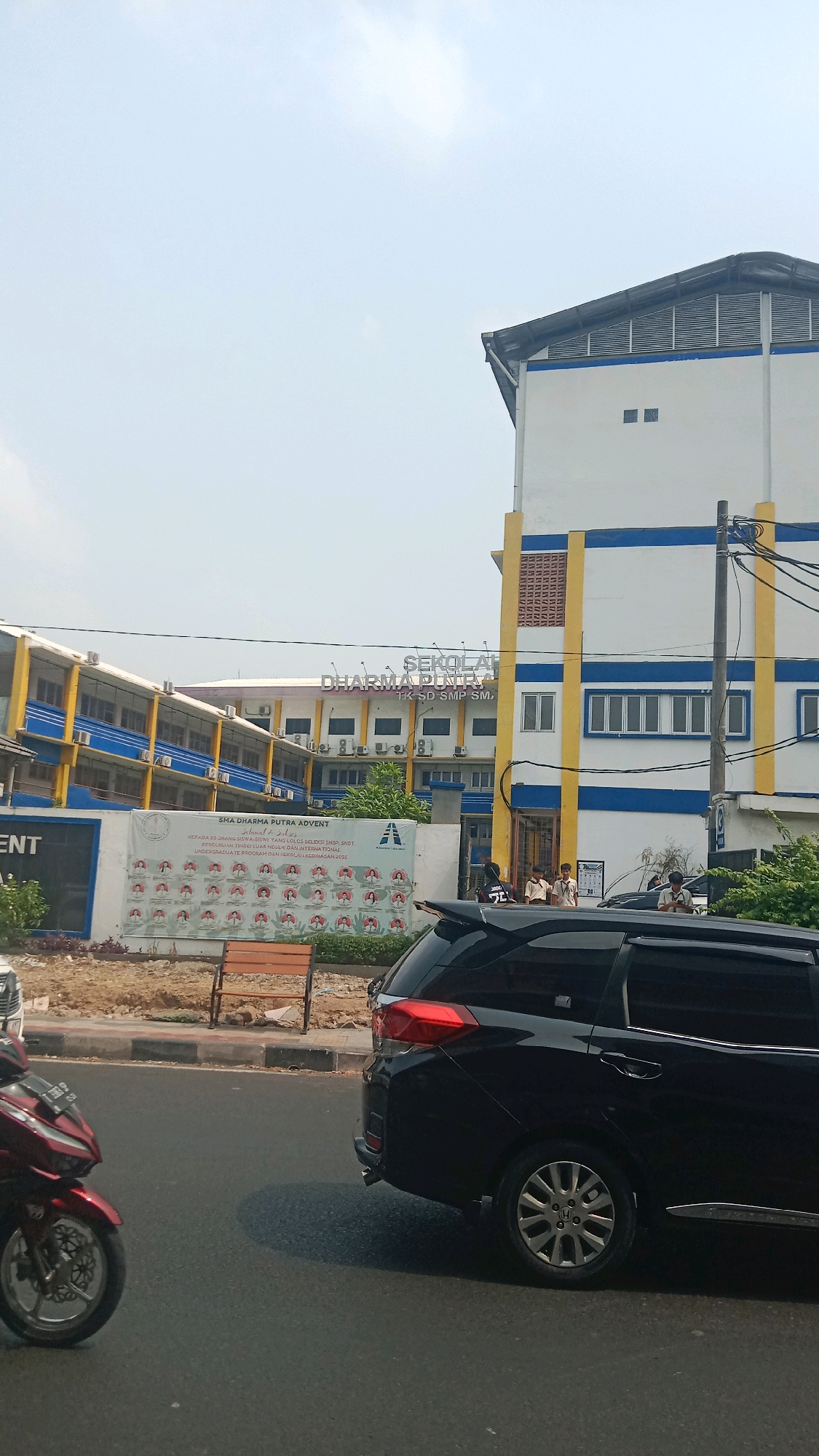 A street scene in front of a school building complex in Bekasi, Indonesia. The school, identified by signage as "SEKOLAH DHARMA PUTRA," features multiple connected buildings with white and blue facades and yellow accents. One building on the right is taller and appears to be a main structure, while a longer building on the left has multiple stories and balconies. A large banner displaying student photos and text related to academic achievement is visible on the front of the school. The sky is bright and overcast. In the foreground, a black SUV is parked on the right side of the road, and a red motorcycle is visible on the left. The road surface is asphalt, and a sidewalk with a wooden bench is present between the road and the school. Several students in uniform are standing near the school entrance. The overall impression is of a typical day at an educational institution in an urban environment.