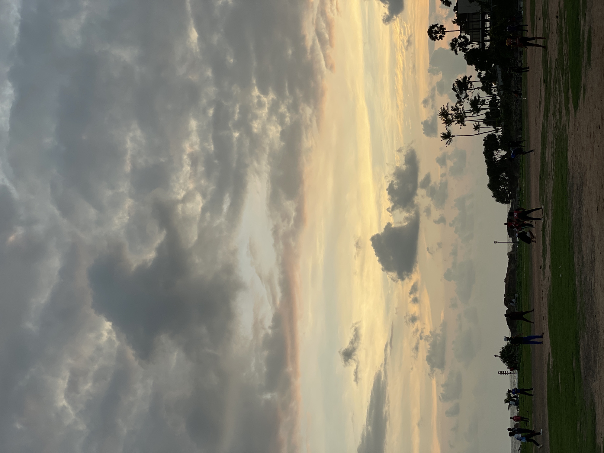 The image depicts a landscape in Galle, Sri Lanka at twilight. The sky is filled with a mix of dark grey and golden clouds. Below the sky is a field where people are scattered, engaging in various activities. Silhouetted palm trees line the horizon. Buildings are visible in the distance.
