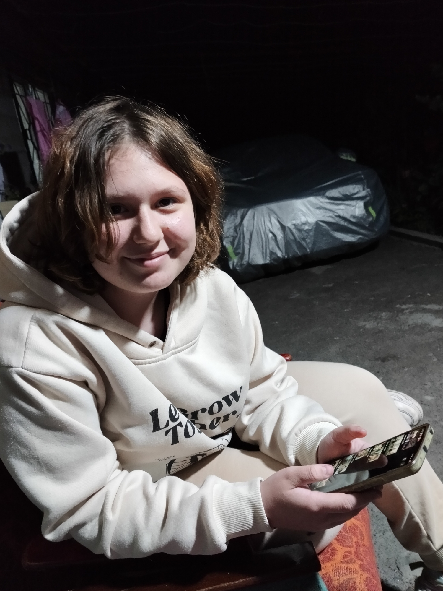 In the dim lighting of an outdoor setting, a young female with shoulder-length brown hair is seated and looking at a smartphone. She is wearing a light-colored hooded sweatshirt with text on the sleeve and light-colored pants. The background shows a dark environment, with a large object covered in a gray tarp visible to her right, suggesting it might be a vehicle. The ground appears to be paved or concrete. The person is holding the smartphone in their hands, with their fingers interacting with the screen. The overall scene is captured at night or in a very low-light condition.