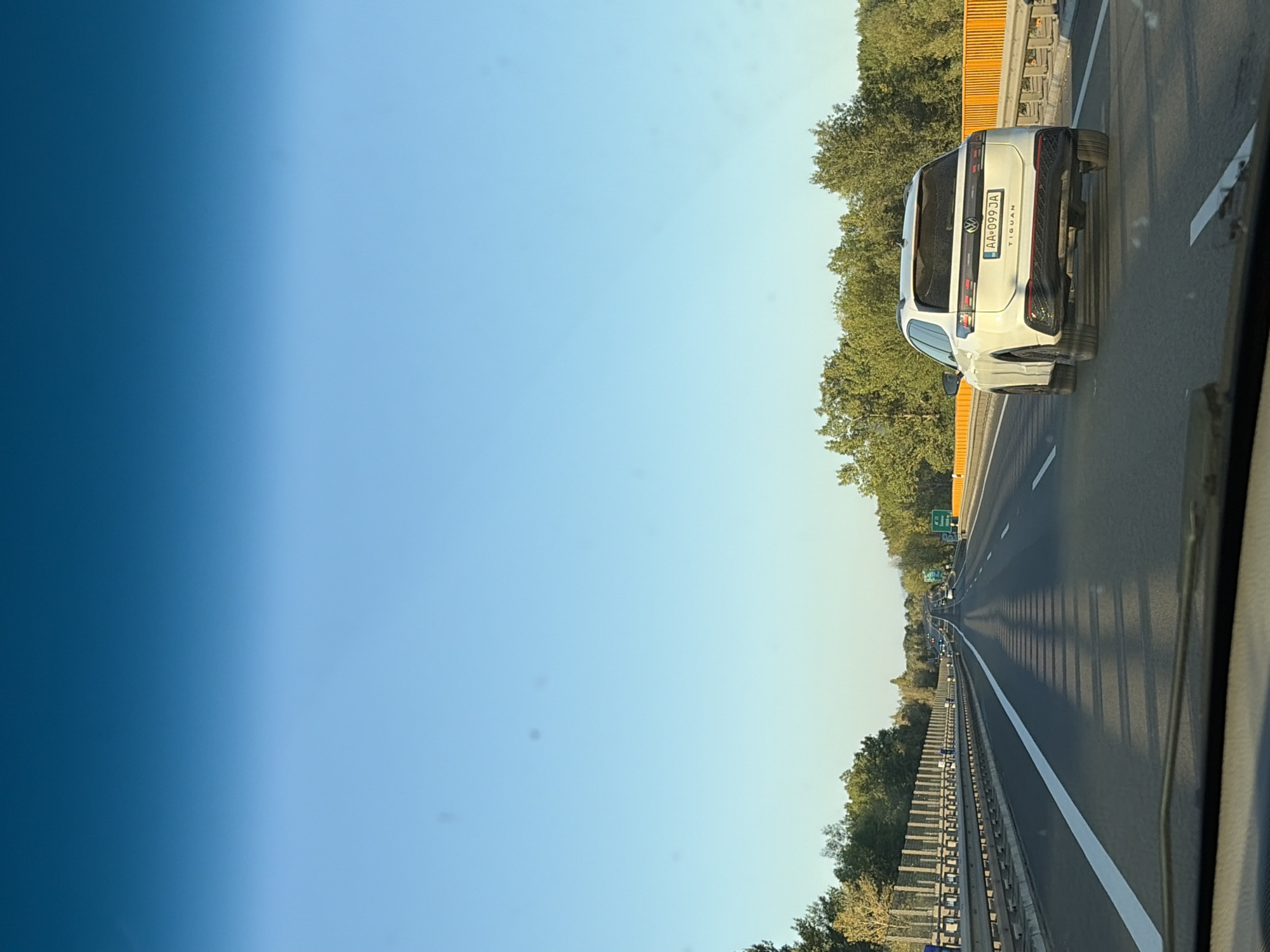 The image shows a white Volkswagen Tiguan driving on a highway. The car has license plate AA-099-JA. The highway has multiple lanes and is bordered by trees on both sides. The sky is clear and blue. The photo appears to have been taken from another vehicle, looking forward. This highway is located in or near the city of Dolná Streda, Slovakia.