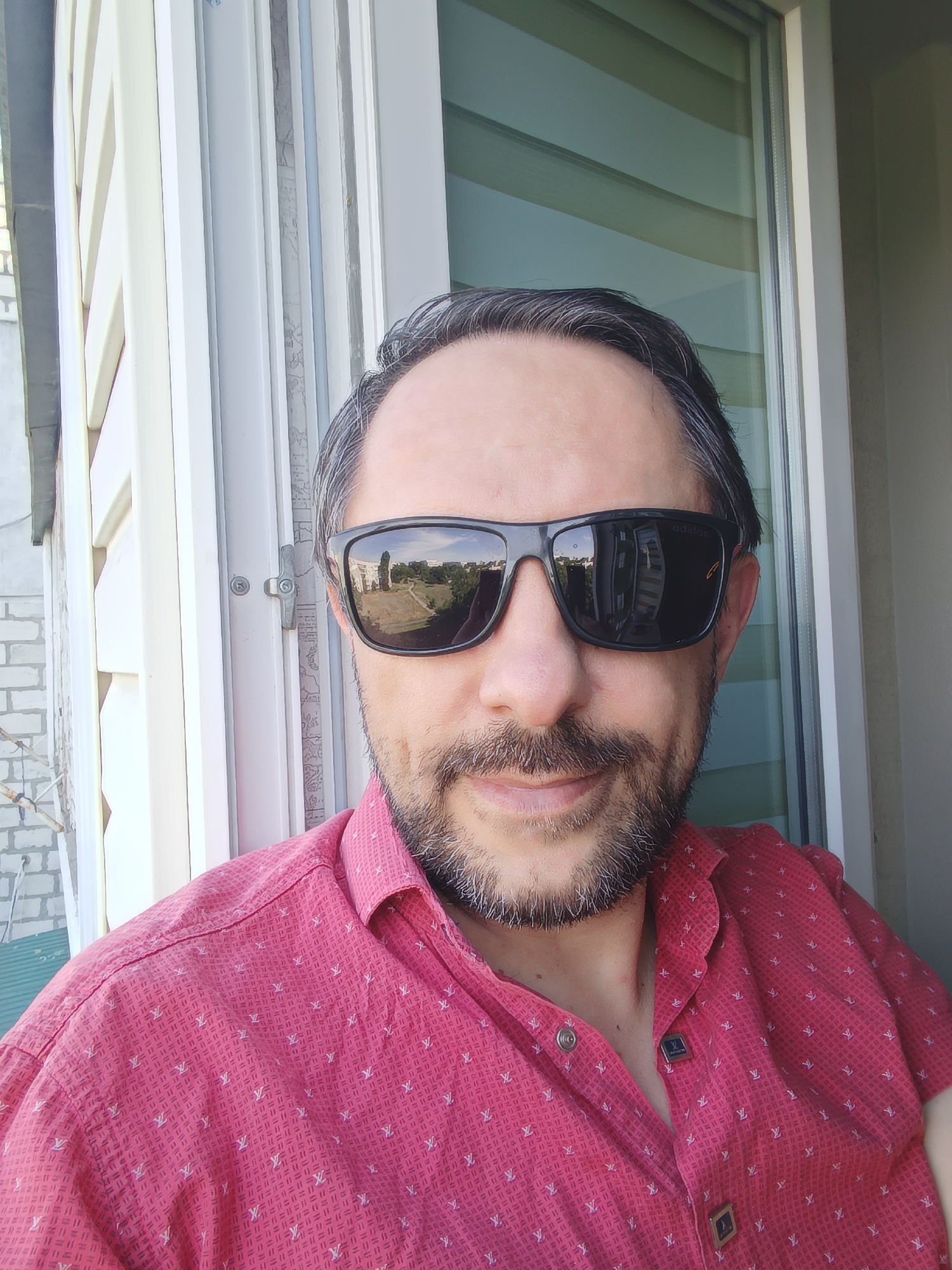 A man with dark hair and a goatee, wearing sunglasses and a pink patterned shirt, poses for a selfie. The background shows a window with blinds and part of a brick building. The man is positioned in front of a window on a balcony. The reflection in his sunglasses shows a cityscape with buildings and trees, consistent with the view from a balcony in Izmail, Ukraine. The lighting suggests it is daytime.