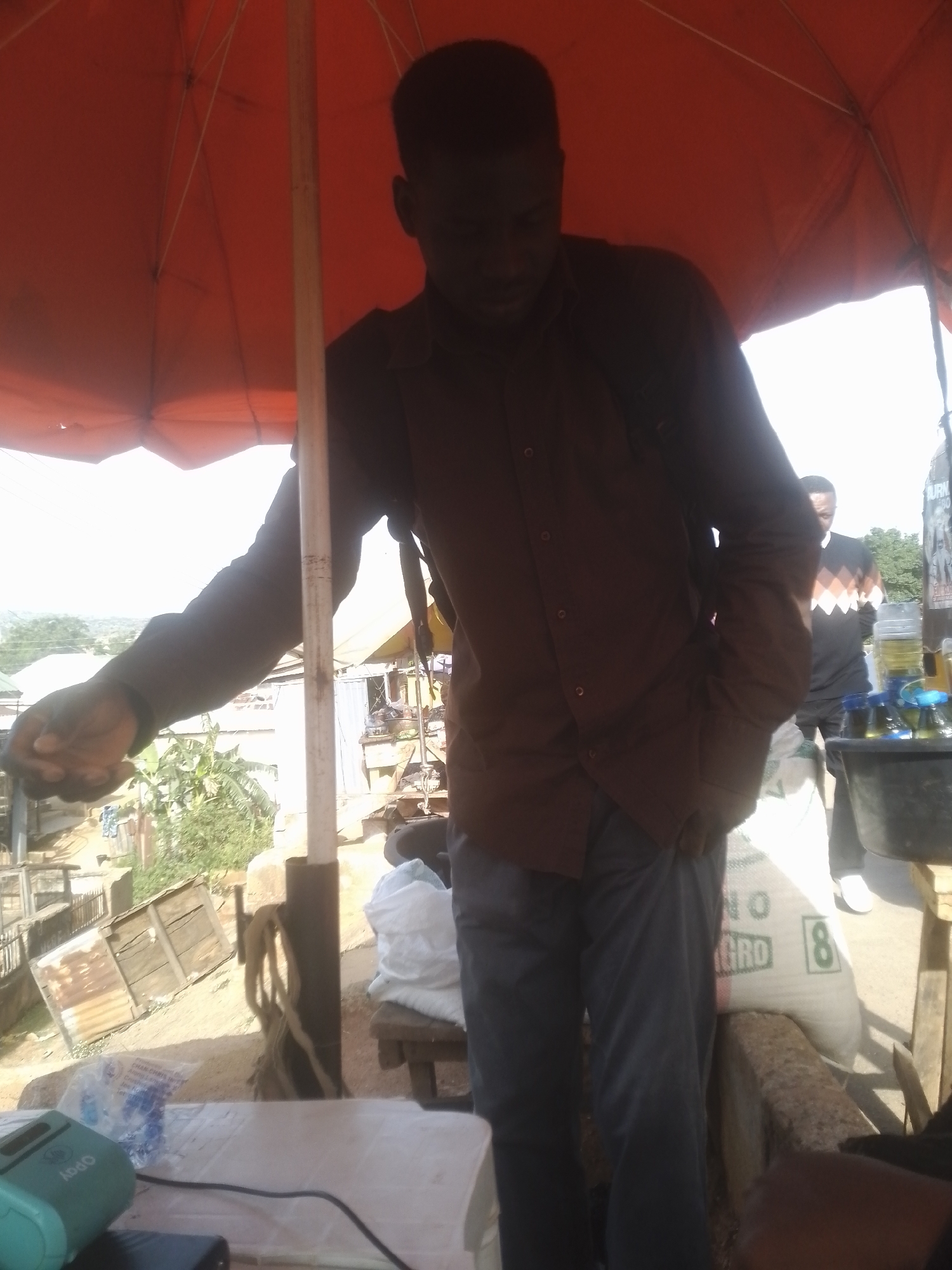 The image shows a medium shot of two men in an outdoor setting, possibly a market. The foreground is dominated by a man wearing a dark brown collared shirt and grey pants. He is partially obscured by an orange umbrella overhead, which casts a shadow. His left arm is extended, and his hand appears to be holding something.
Behind him, another man is visible, wearing a dark sweater with a diamond pattern in orange and white. He is looking down, and his face is not clearly visible.
The scene appears to be a busy, perhaps somewhat rustic, environment. In the background, there are buildings with corrugated metal roofs and some vegetation. There is also a large white sack with green lettering visible. The lighting suggests it is daytime, possibly sunny, given the bright sky.
On a table in the foreground, several items are visible, including a light blue electronic device with some branding. A black cable is also present. To the right, there are several bottles containing yellow liquid, suggesting a vendor stall.
The overall impression is of a casual, everyday scene taking place outdoors. The focus is on the two individuals and the goods around them. There is no clear indication of a specific event or emotional state.