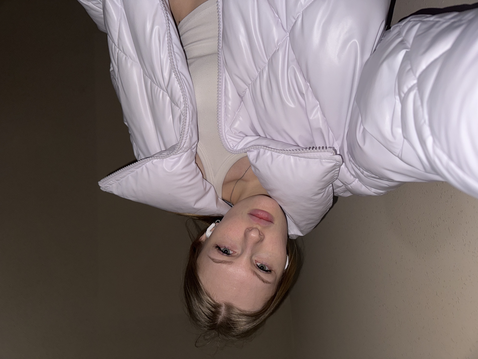 The original image presents a close-up, inverted (upside-down) view of a young woman looking directly at the camera. She has light blonde or light brown hair, fair skin, and striking blue-green eyes. Her lips are slightly parted, conveying a neutral or slightly contemplative expression.
She is dressed in a shiny, light purple (lilac) quilted puffer jacket, which is partially unzipped, revealing a light beige or cream ribbed top underneath. A thin silver necklace with a small pendant is visible on her chest. White wireless earbuds are worn in both ears.
The background is an undifferentiated, dark greyish-brown wall or surface, suggesting an indoor setting or a poorly lit environment. The lighting, possibly artificial, highlights her face and the glossy texture of her jacket. There is no visible text in the image. The scene appears to be a self-portrait or selfie taken in a casual setting, possibly in Perm, Russia, as per the provided context.