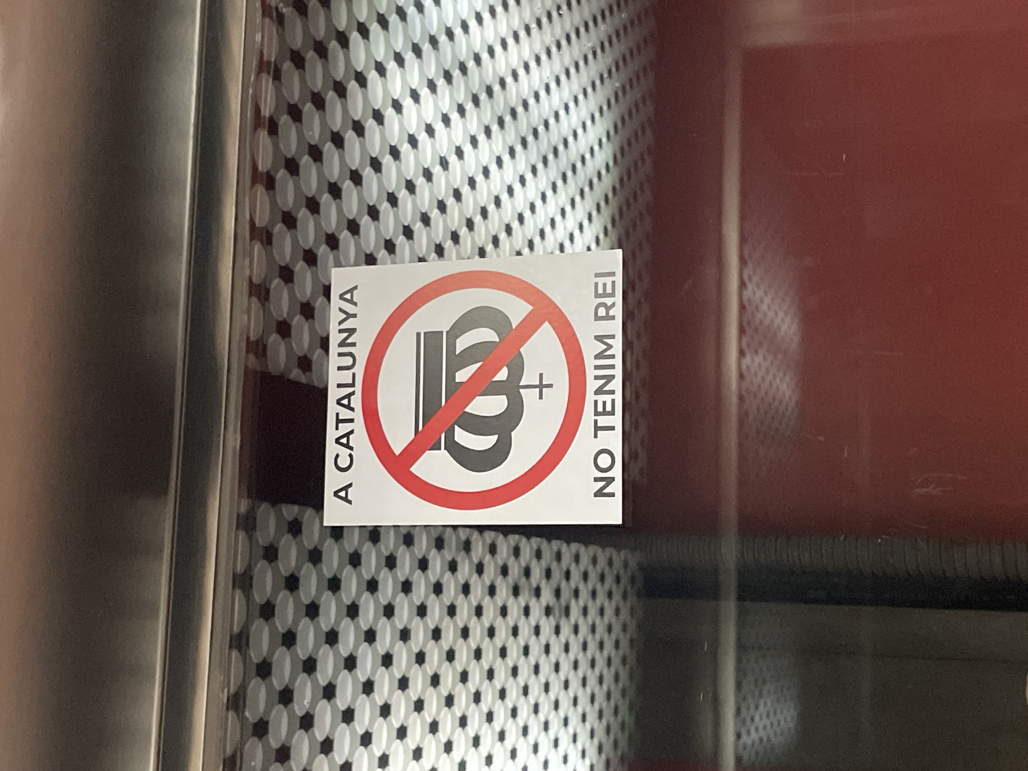A sticker is affixed to a textured surface, likely a window or wall, featuring a design of a crown crossed out with a red circle and diagonal line. The text “A CATALUNYA” is on the left side of the sticker, and “NO TENIM REI” is on the right. The phrase translates to "In Catalonia, we don't have a king" or "We don't have a king in Catalonia".